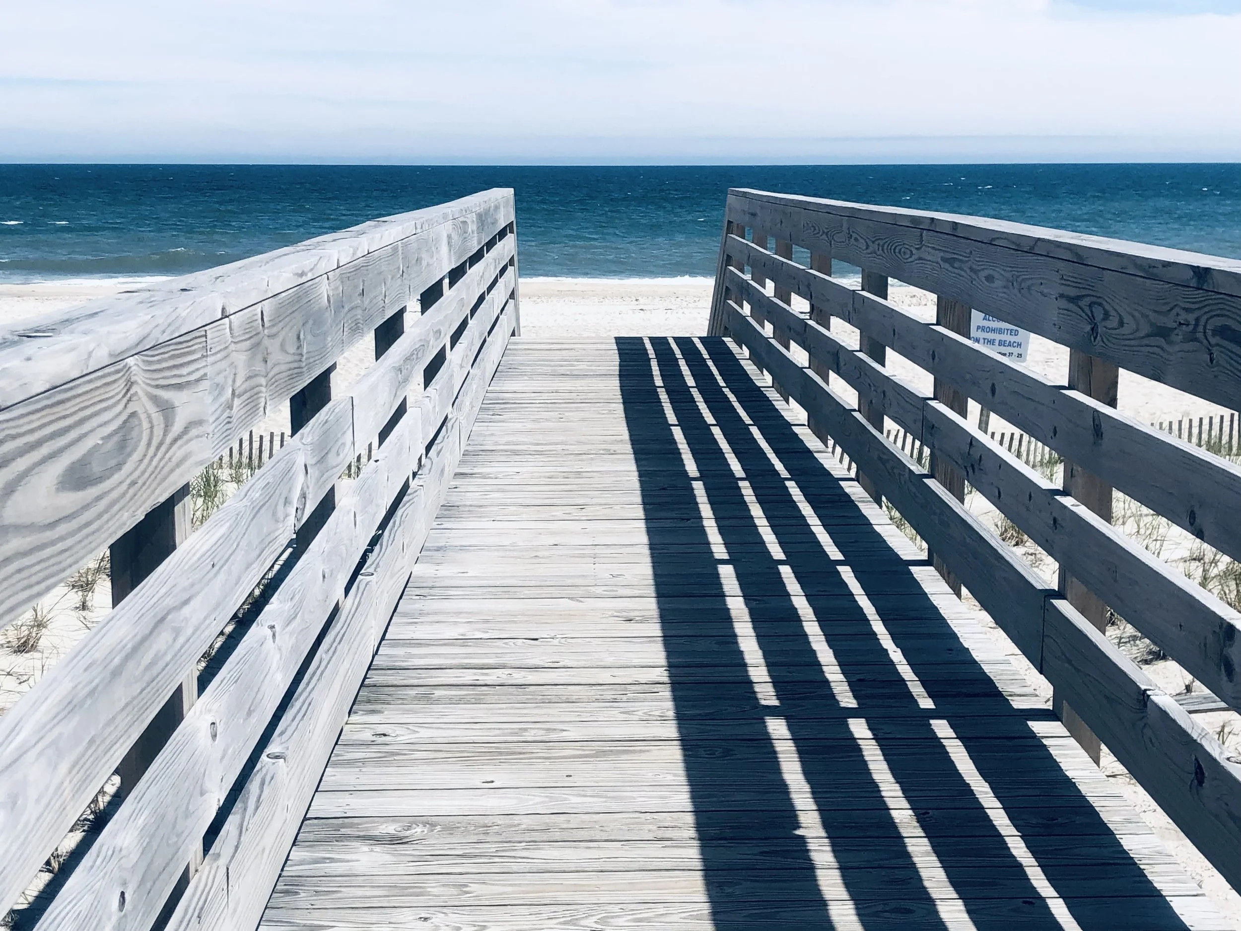 A wooden boardwalk extending over sand dunes toward the ocean with waves and a cloudy sky in the background.