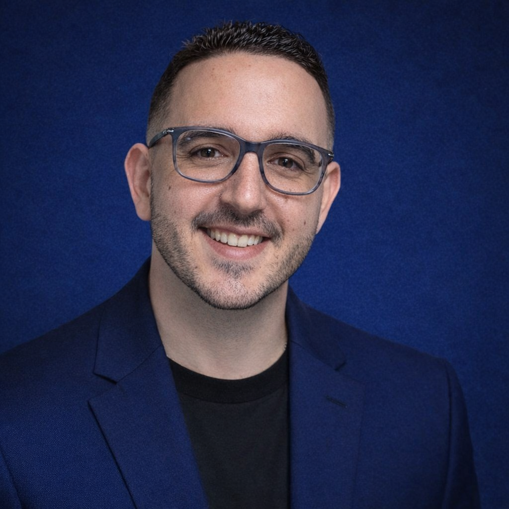 A man with short dark hair, wearing glasses, a dark blazer, and a black shirt, smiling against a solid dark blue background.