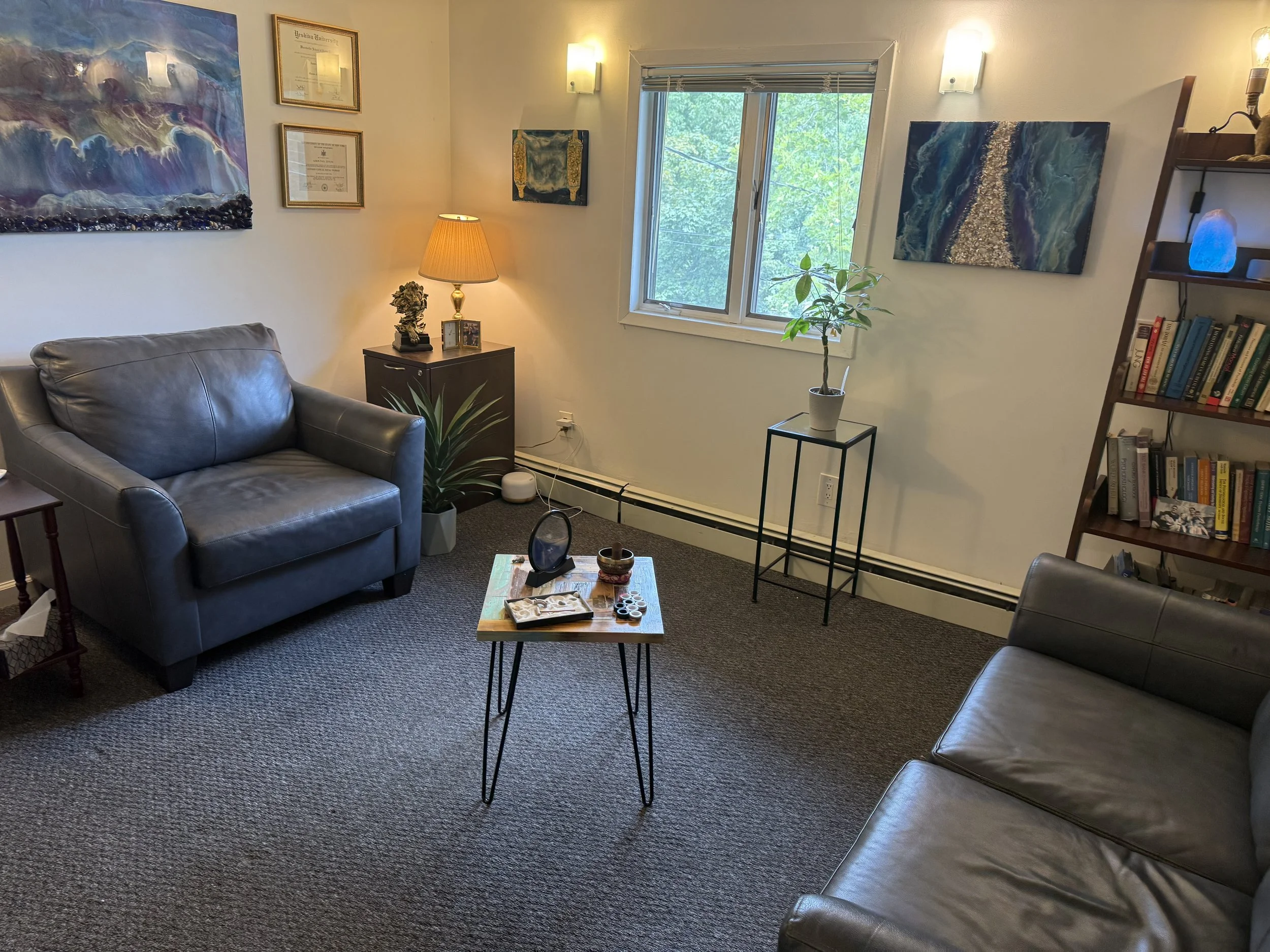 A cozy living room corner with two leather sofas, wall art, a small wooden table, a lamp, a mini bookshelf, and a window with trees outside.