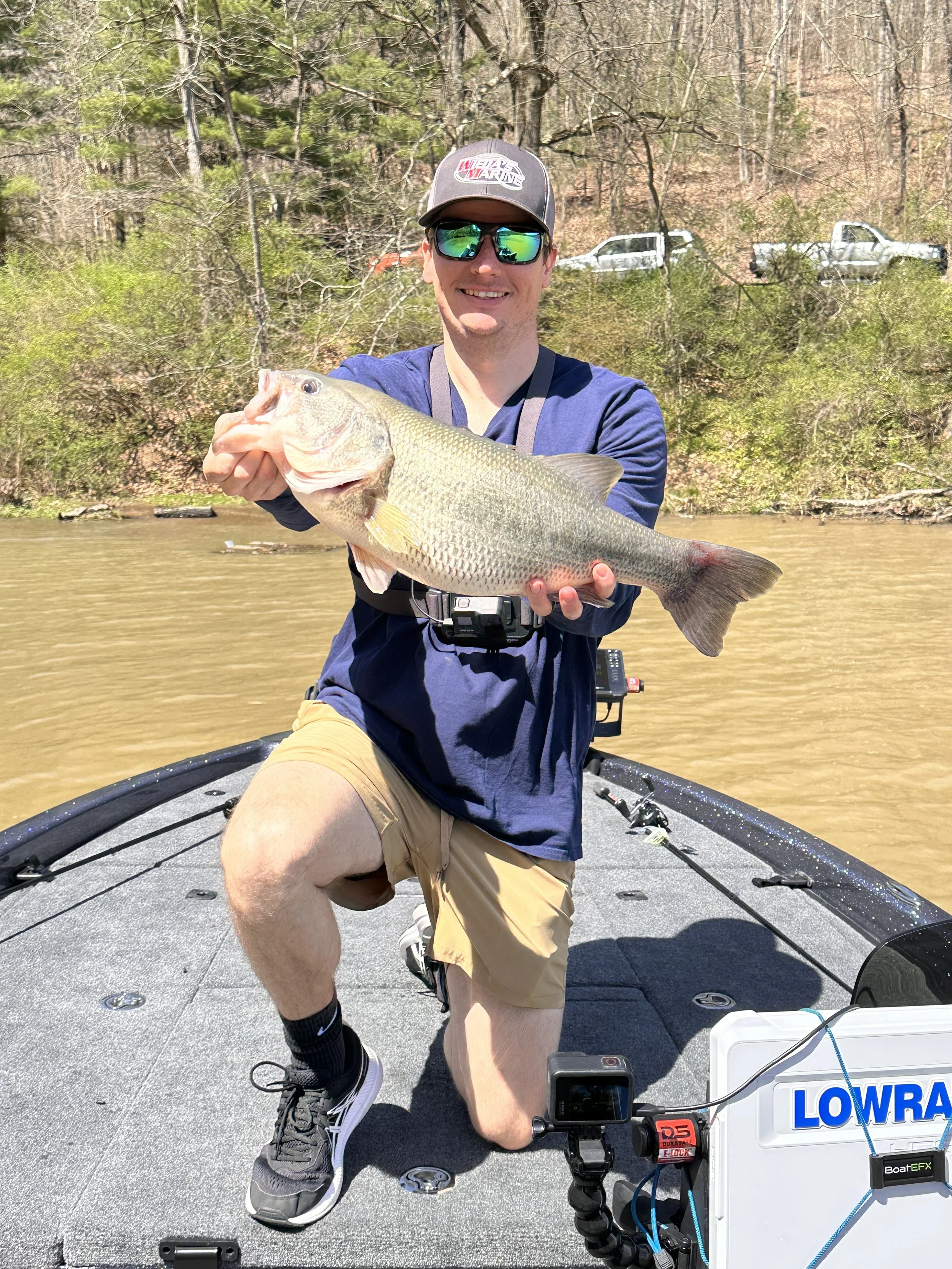 A man in sunglasses and a cap kneels on a boat, holding a large fish he caught, with a river and wooded shoreline in the background.