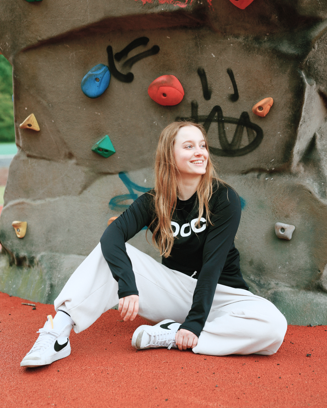A young woman with long, wavy red hair sitting on a red rubberized surface in front of a rock climbing wall with colorful holds and graffiti.