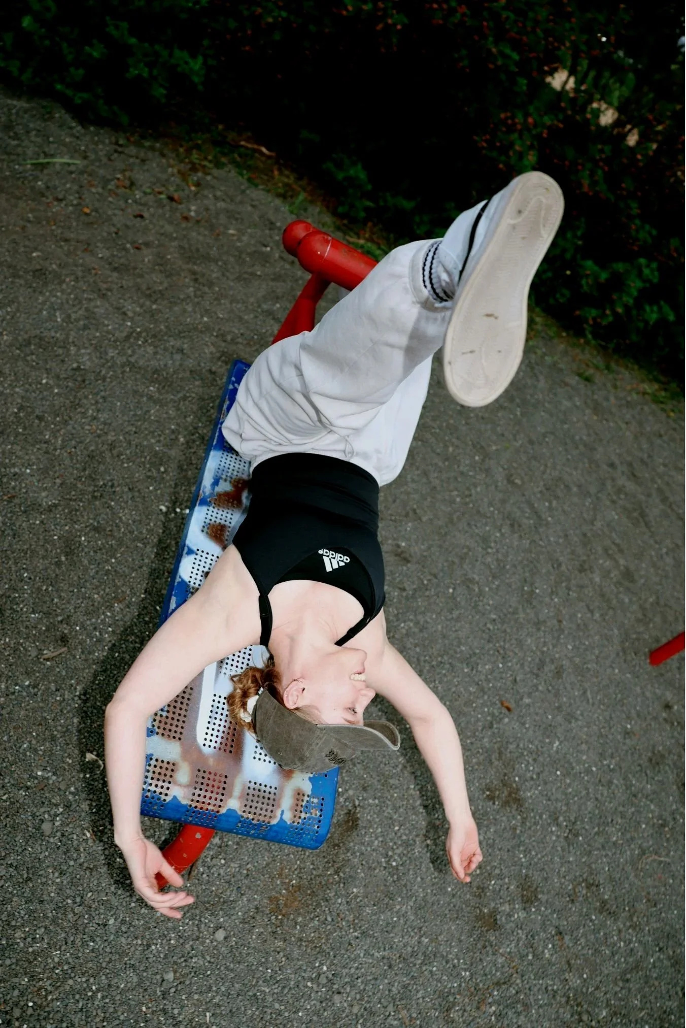 A young woman with red hair lying on a small outdoor sledge on gravel, smiling with her arms stretched out and wearing a tan baseball cap, black tank top, and shorts, with her eyes closed.