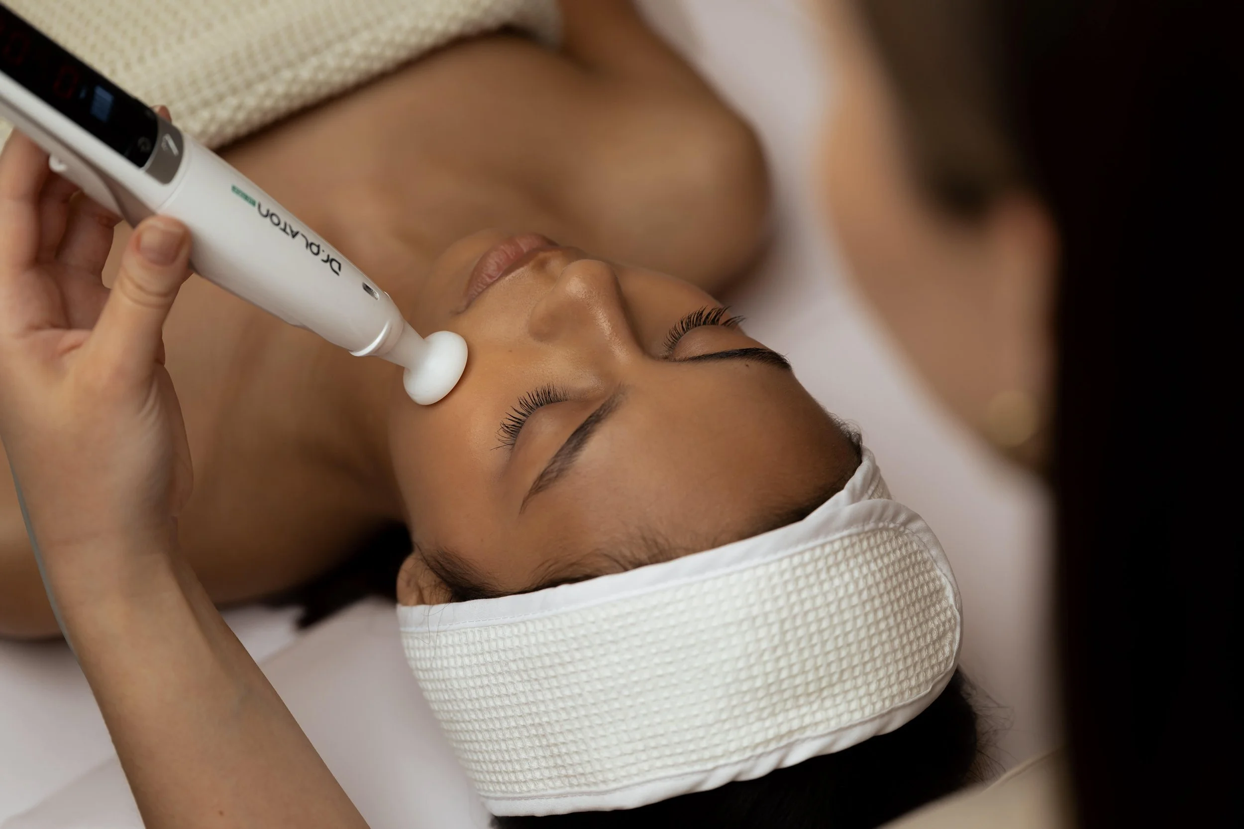 A woman lying on her back with eyes closed, receiving a facial treatment with a white dermal oxygen device on her face, in a spa or clinic setting.