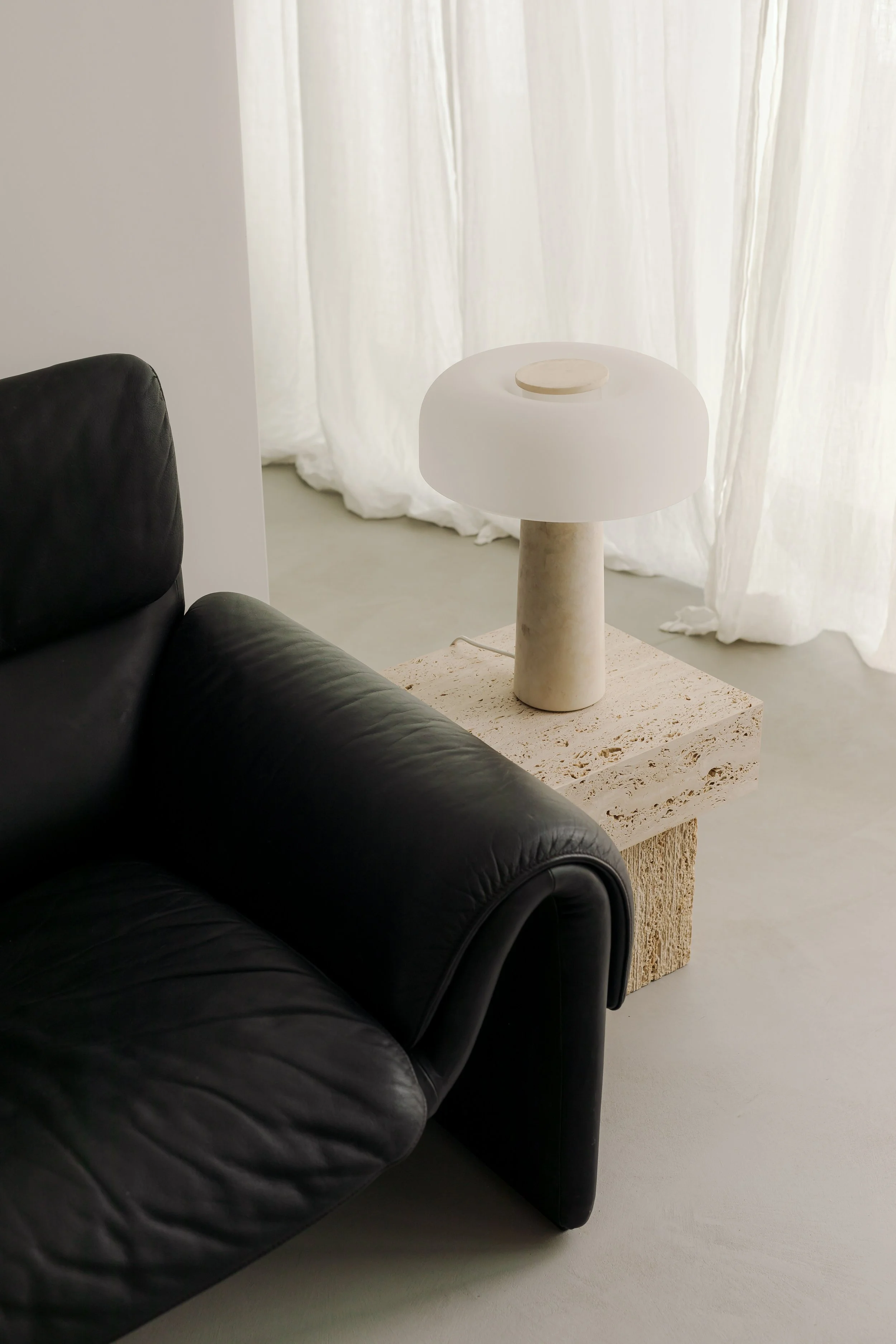 A black leather sofa in the foreground, with a beige textured side table holding a modern white lamp with a cylindrical base and a rounded shade. Behind the lamp, there are sheer white curtains hanging in front of a window, allowing light to softly diffuse into the space.