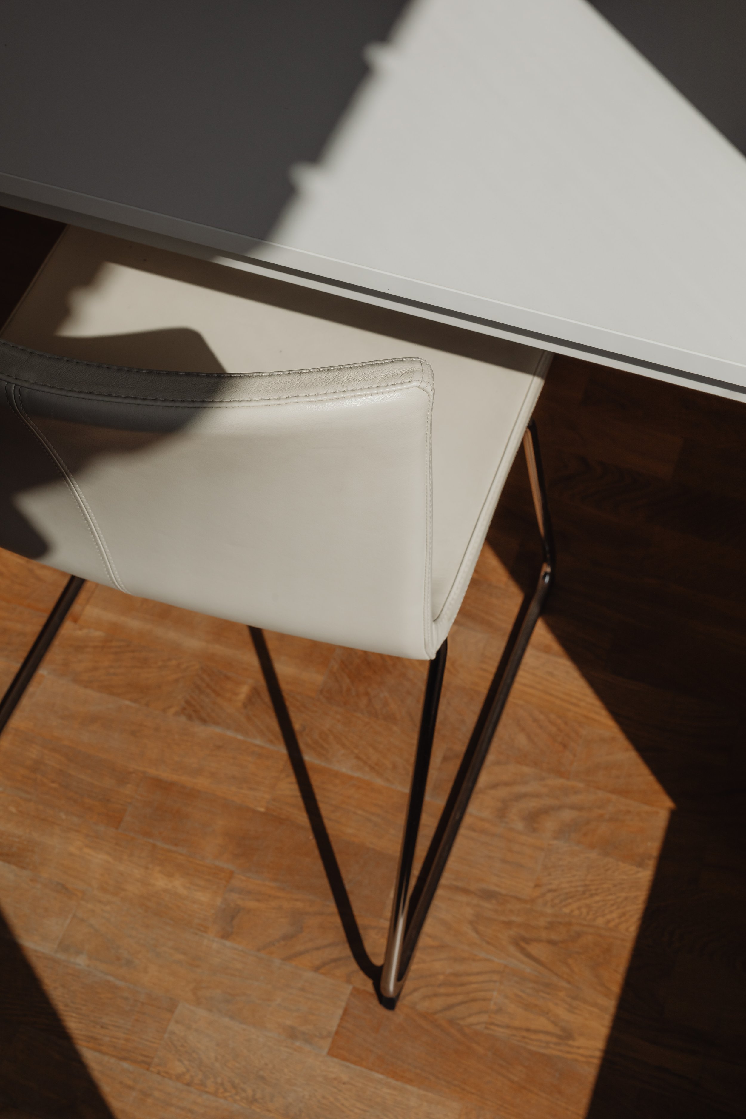 A white leather chair with thin black metal legs is positioned on a wooden floor, partially shadowed.