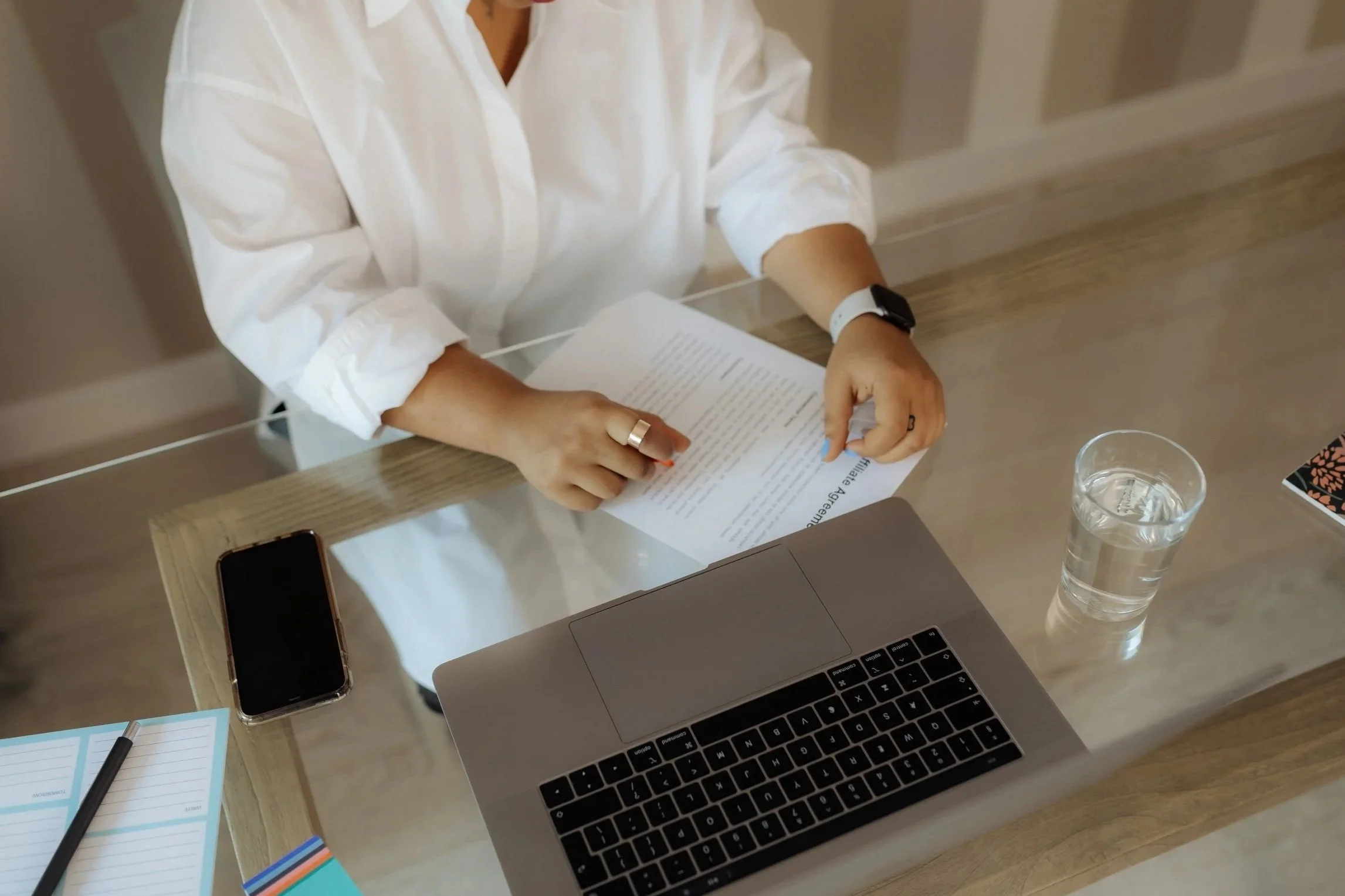 A person in a white shirt sitting at a wooden desk with a laptop, a smartphone, a glass of water, and writing utensils. The person is holding a document and wearing a watch and a ring.