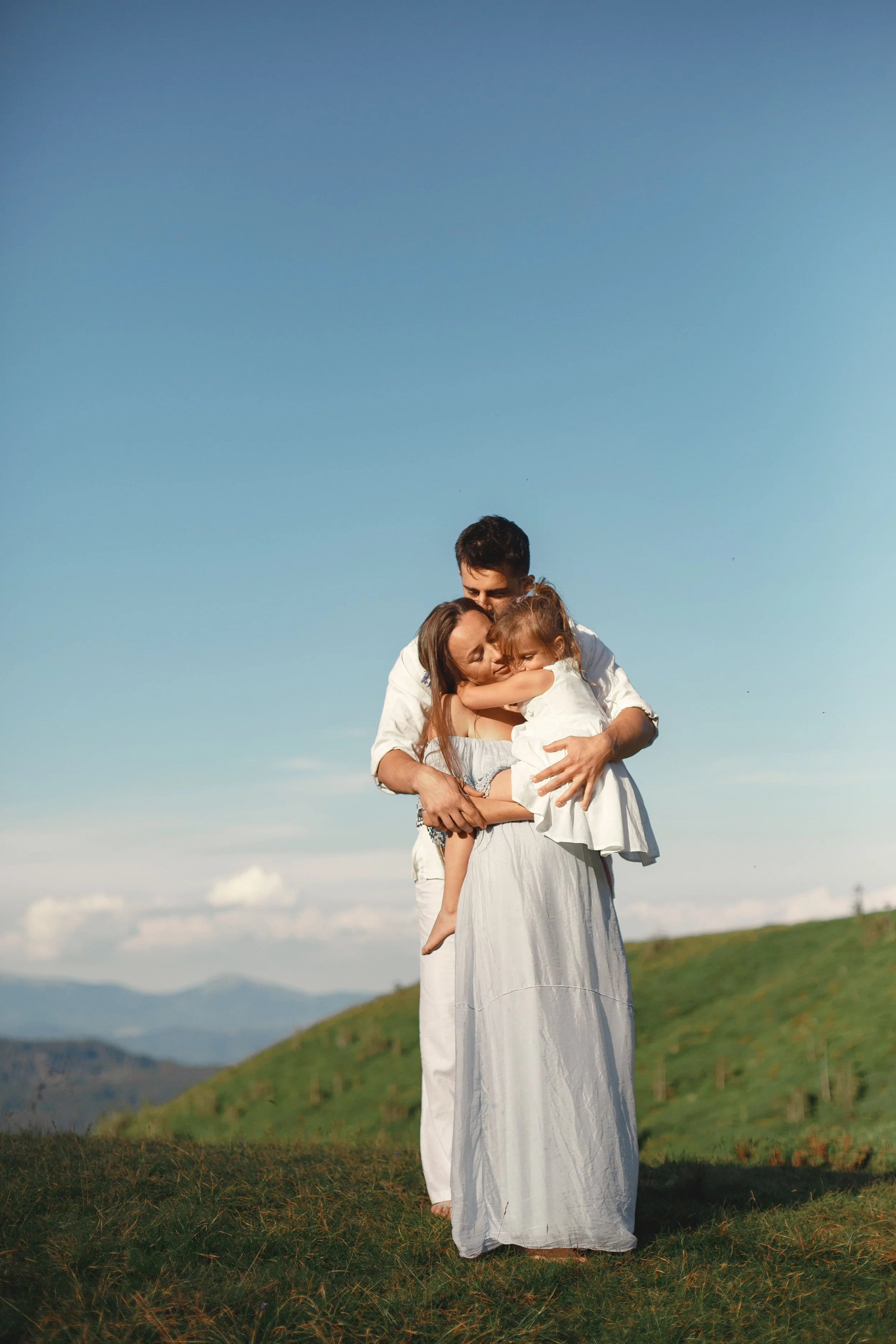 Family of three, a man, woman, and young girl, embracing in a grassy outdoor field under a clear blue sky with rolling hills in the background.