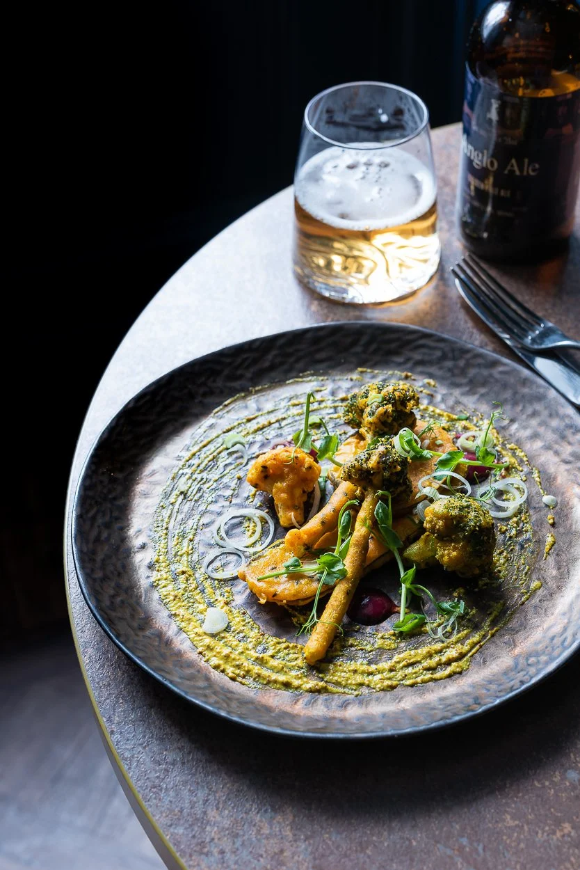 A black plate with assorted tempura vegetables, garnished with microgreens and sliced onions, on a white table. There is a glass of beer and a bottle labeled "Anglo Ale" in the background.