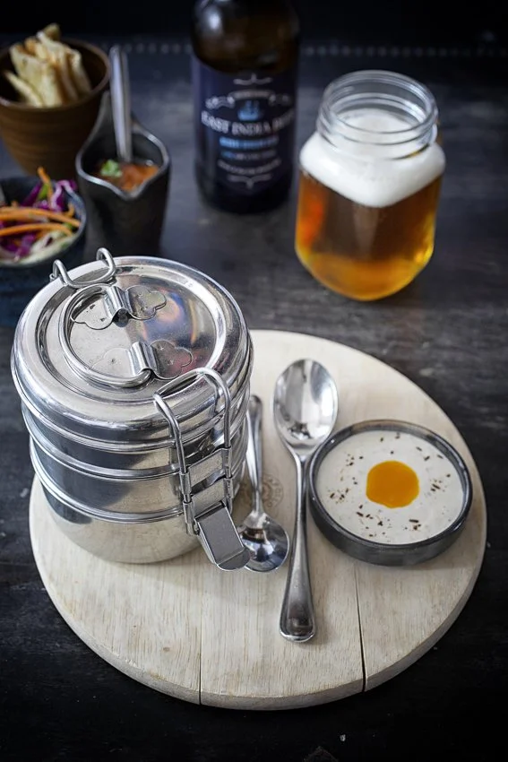 A wooden serving board with a metal food cover, a spoon, a small dish of creamy egg yolk, and a glass of beer on a dark table. In the background, there is a jar of beer, a bottle, and small bowls of salad and dipping sauce.