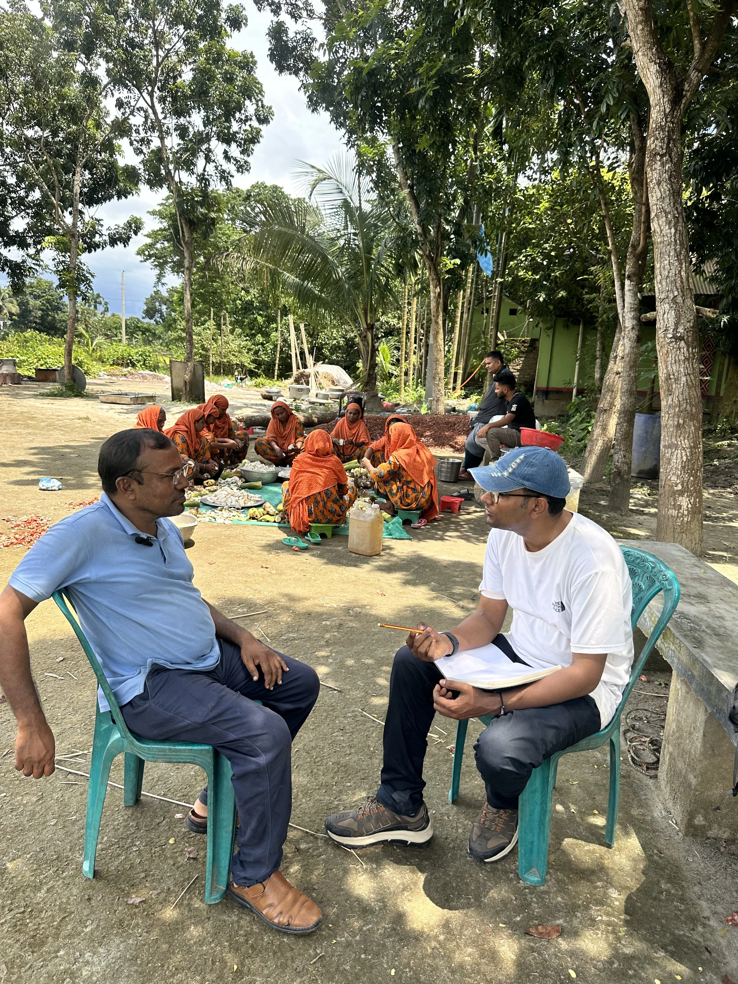 Two men are seated on blue plastic chairs having a conversation outdoors, while a group of women dressed in traditional attire sit cross-legged on the ground nearby, possibly involved in a communal activity or prayer, with a backdrop of trees and greenery.
