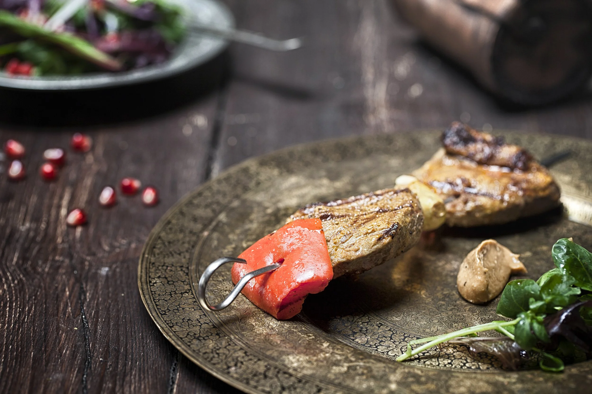 Grilled steak with red bell pepper on a decorative plate, with greens and a side dish in the background.