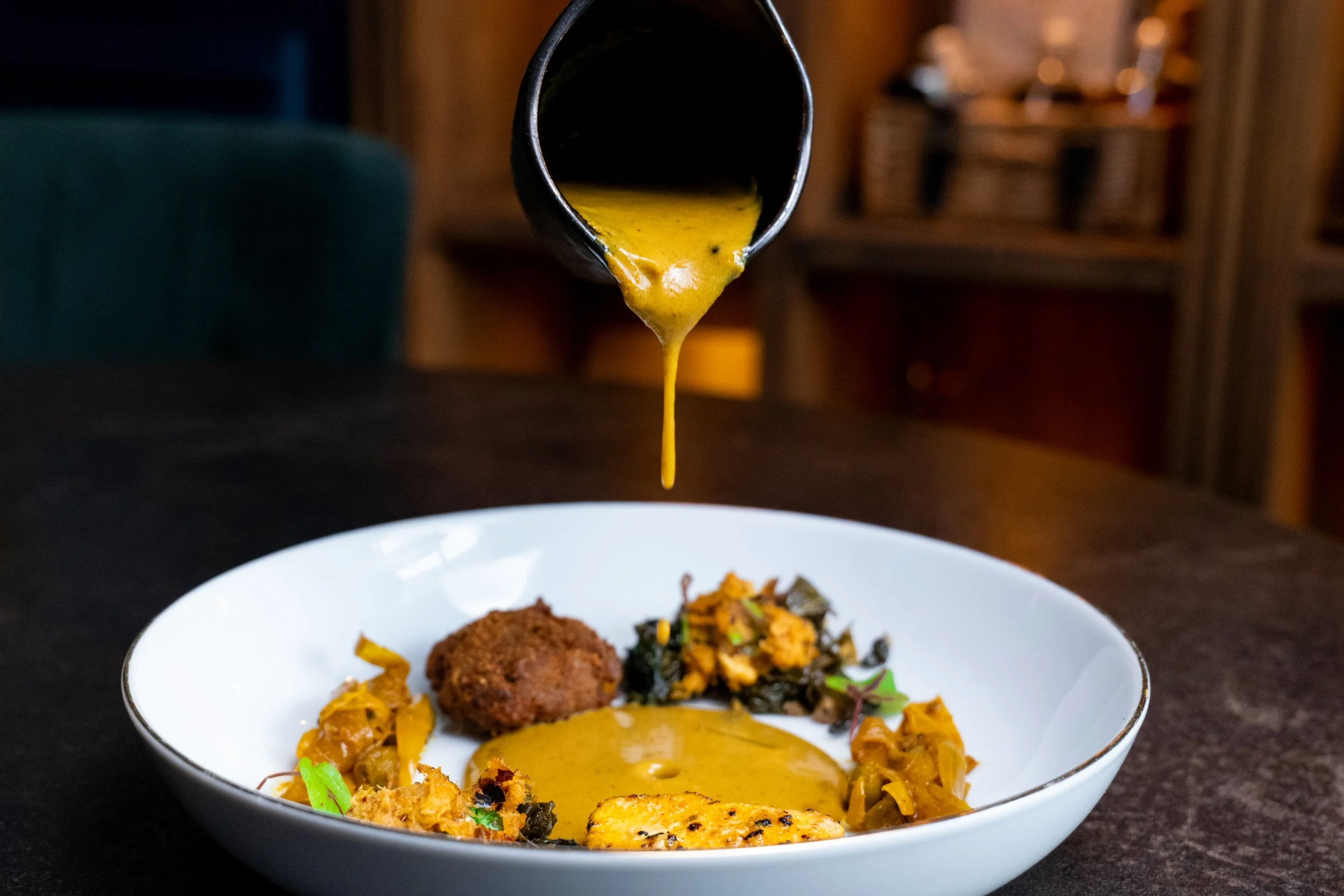 A black ladle pouring yellow curry over a bowl of rice and vegetables, with some fried items on the side in a white bowl.