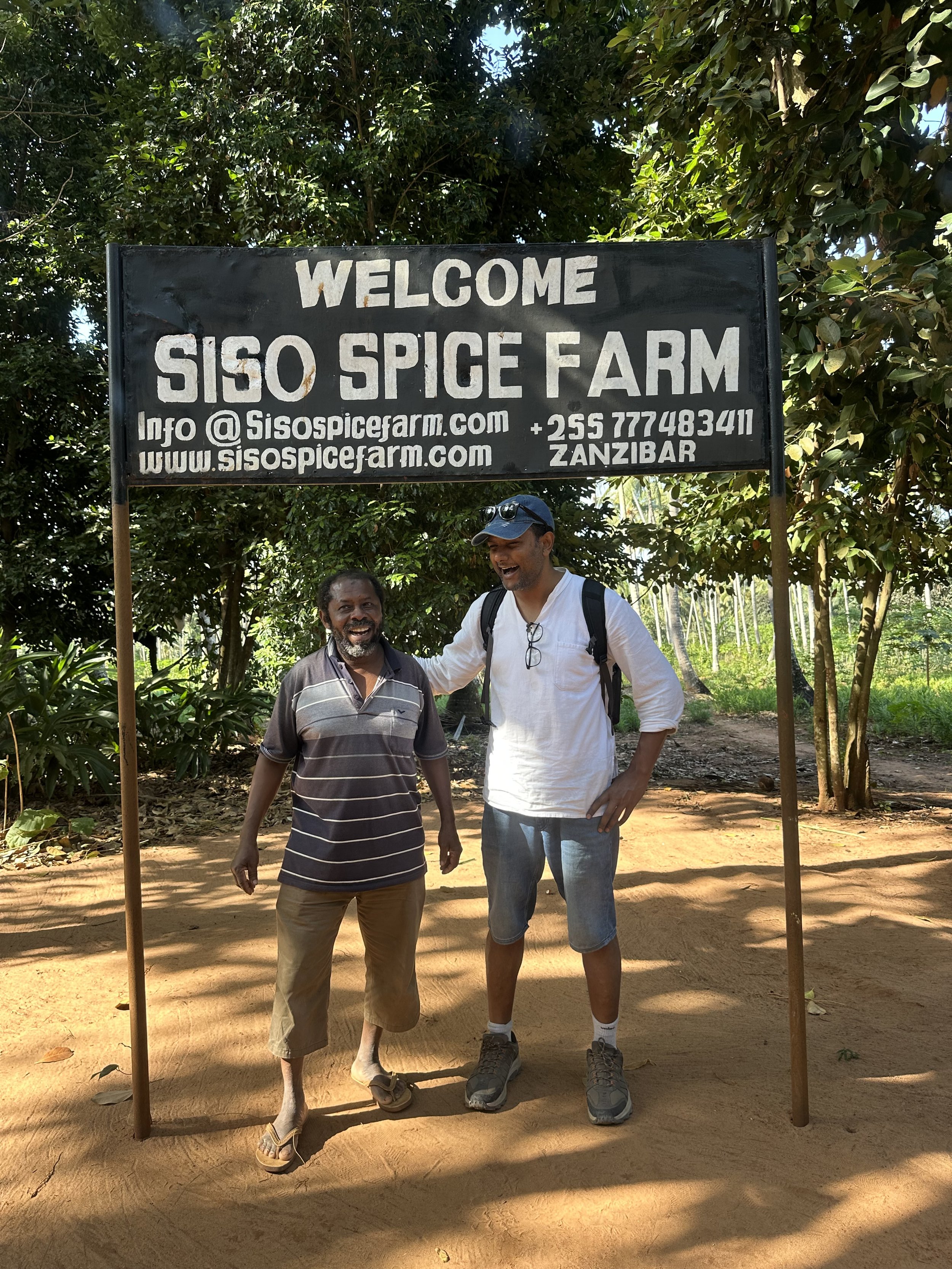 Two men smiling and standing together under a sign that says "Welcome SISO Spice Farm" in Zanzibar, surrounded by trees and a dirt pathway.