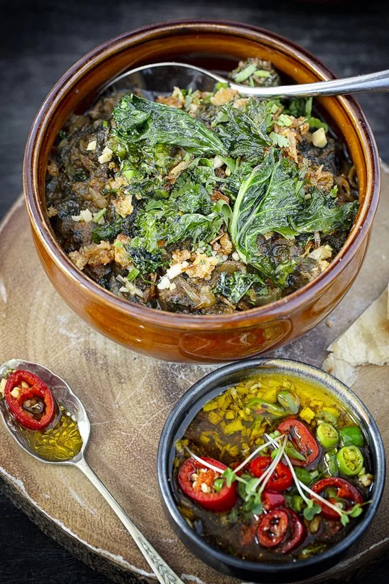 A bowl of cooked greens topped with herbs, next to a small bowl of chopped peppers in oil, on a wooden serving board.