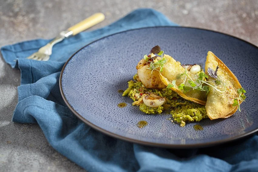 Gourmet dish with fried elements and microgreens on a textured blue plate, placed on a blue cloth napkin with a fork.