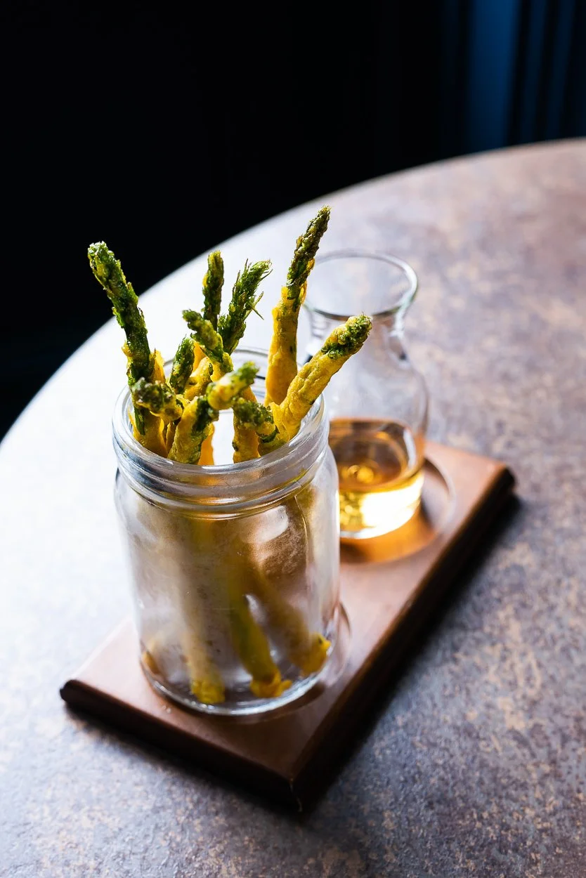 Fried green asparagus spears served in a glass jar on a wooden platter with a small pitcher of dipping sauce in the background.