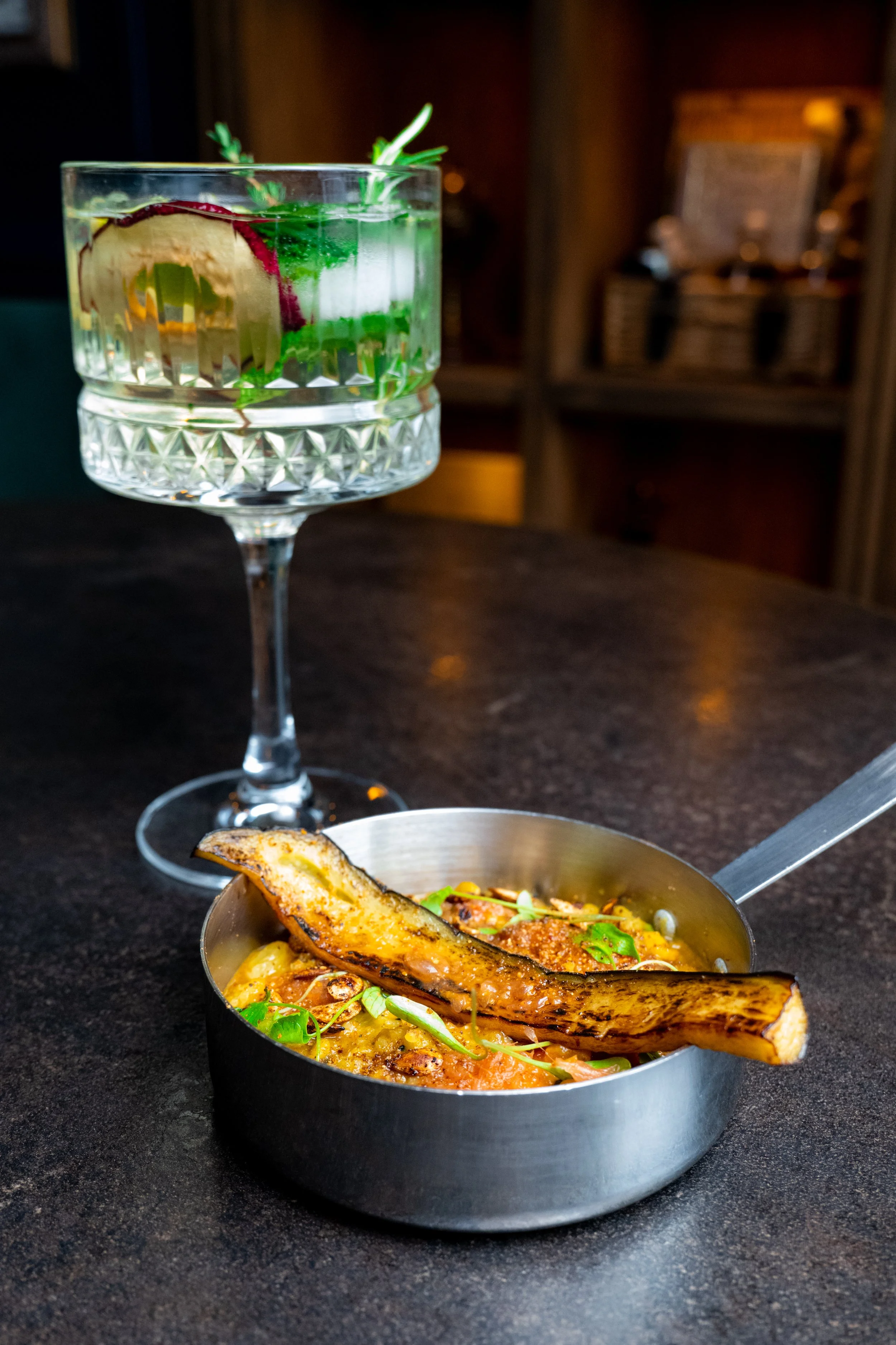 A cocktail glass filled with a clear drink, garnished with a lemon wedge, and a sprig of herbs, sits on a dark table. A metal bowl with food and a piece of grilled fish is partially visible in front of the glass.