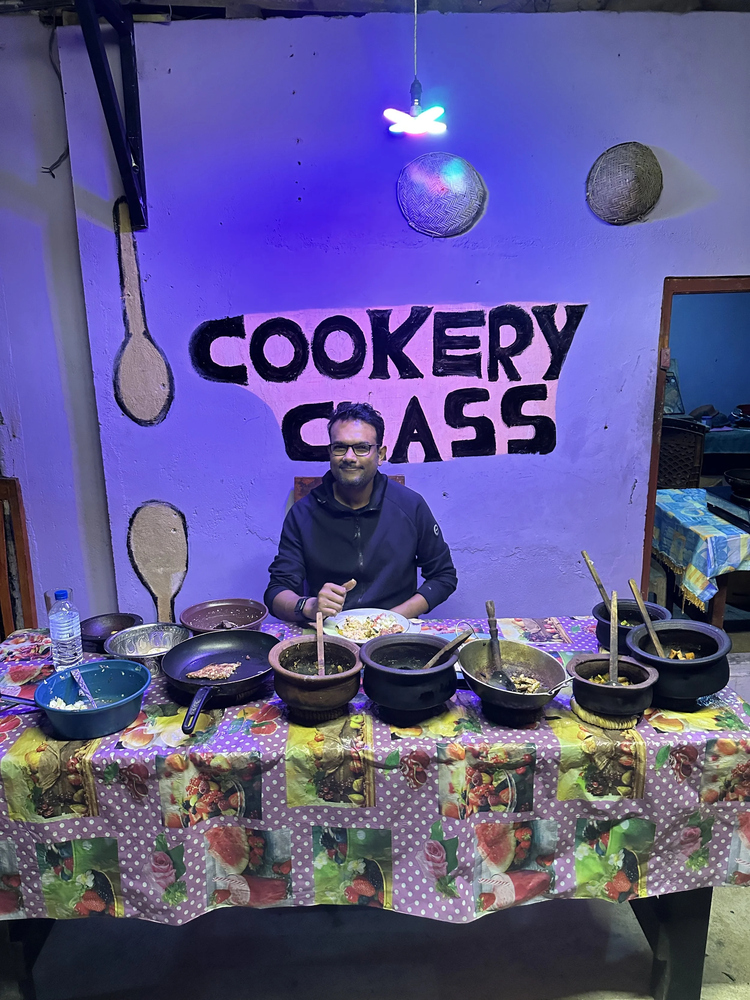 A man sitting at a decorated table with various bowls of food, in front of a wall with the sign 'Cookery Class' painted on it. The wall has two woven baskets and a colorful light fixture hanging above.