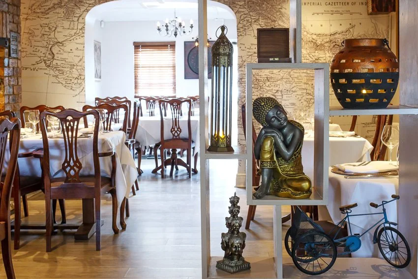 Dining room with wooden tables and chairs, decorative artifacts including a statue, lantern, large vase, and a miniature bicycle on a white shelf, with a map-patterned wall and chandelier.