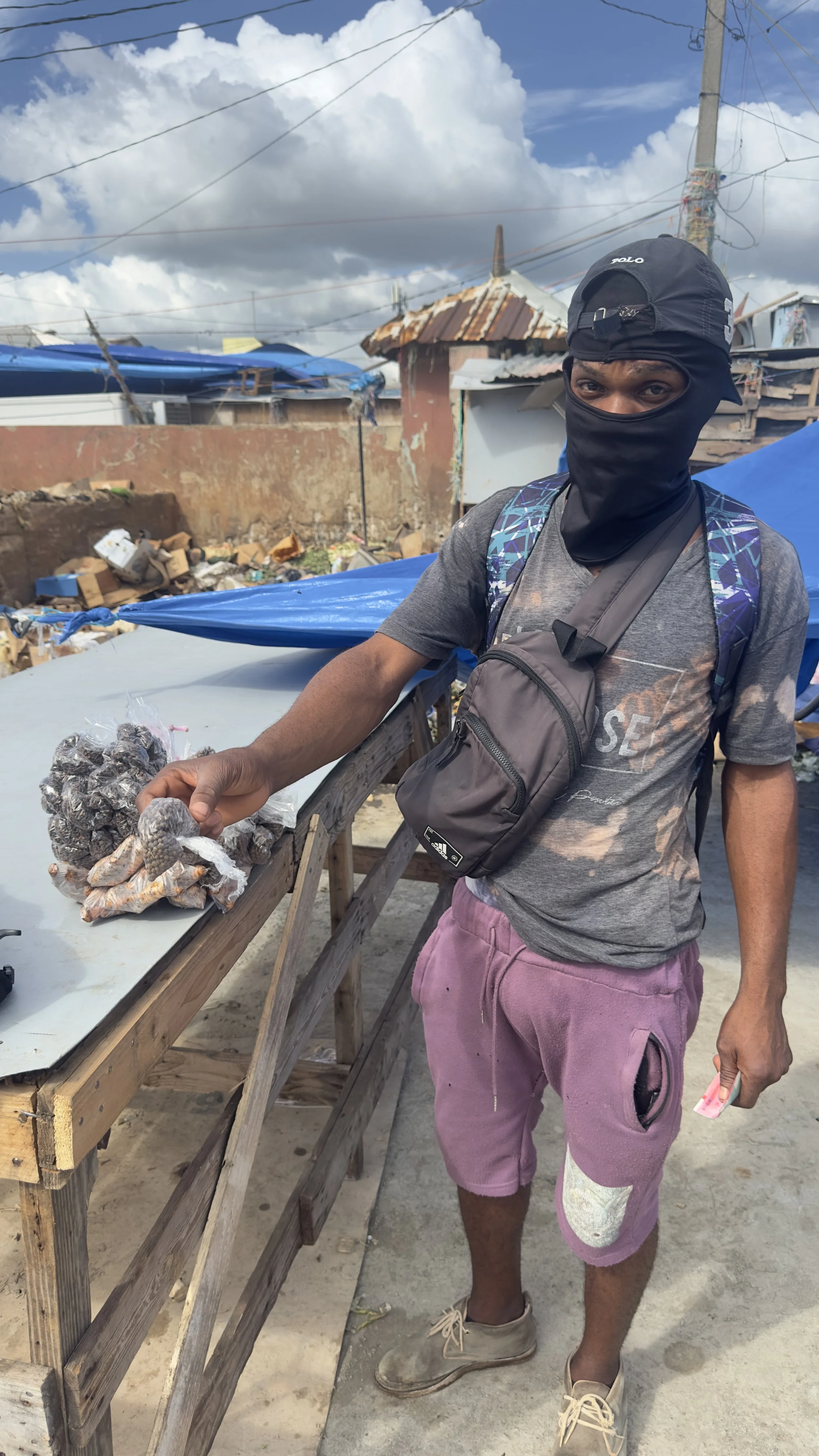 A young man wearing a black face mask, grey t-shirt, pink shorts with a white patch, and tan shoes, standing outdoors in a makeshift market area. He is holding a bag of what appears to be dried goods or snacks, and has a backpack over one shoulder. Behind him, there are wooden tables, blue tarps, and makeshift buildings with rusty roofs, under a partly cloudy sky.