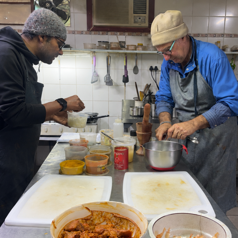 Two men cooking in a kitchen, surrounded by various containers of spices and ingredients, with one chopping and the other preparing food.