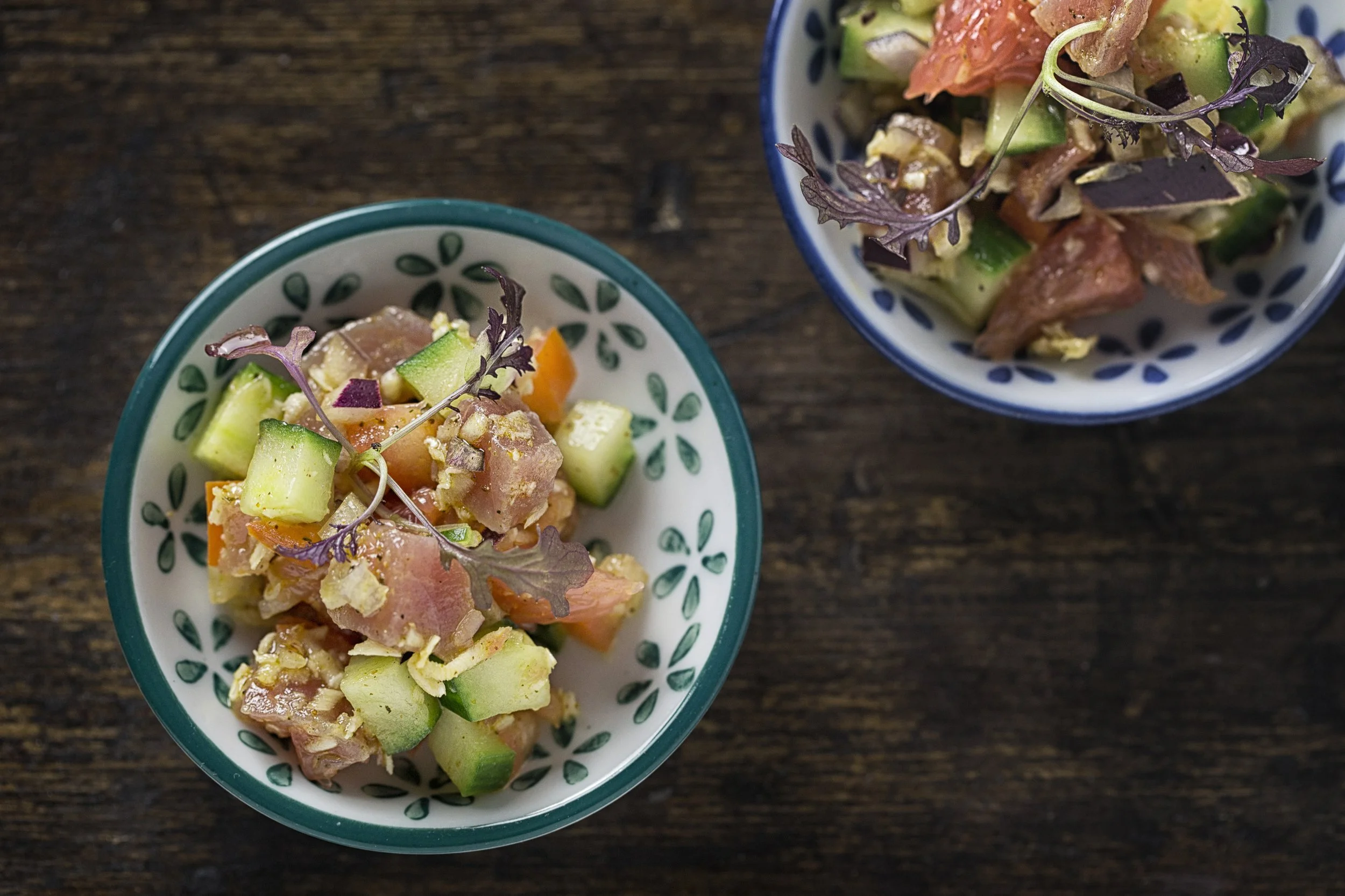 Two bowls of poke salad on a wooden surface. The bowls contain chopped cucumbers, carrots, and fish, garnished with purple microgreens.