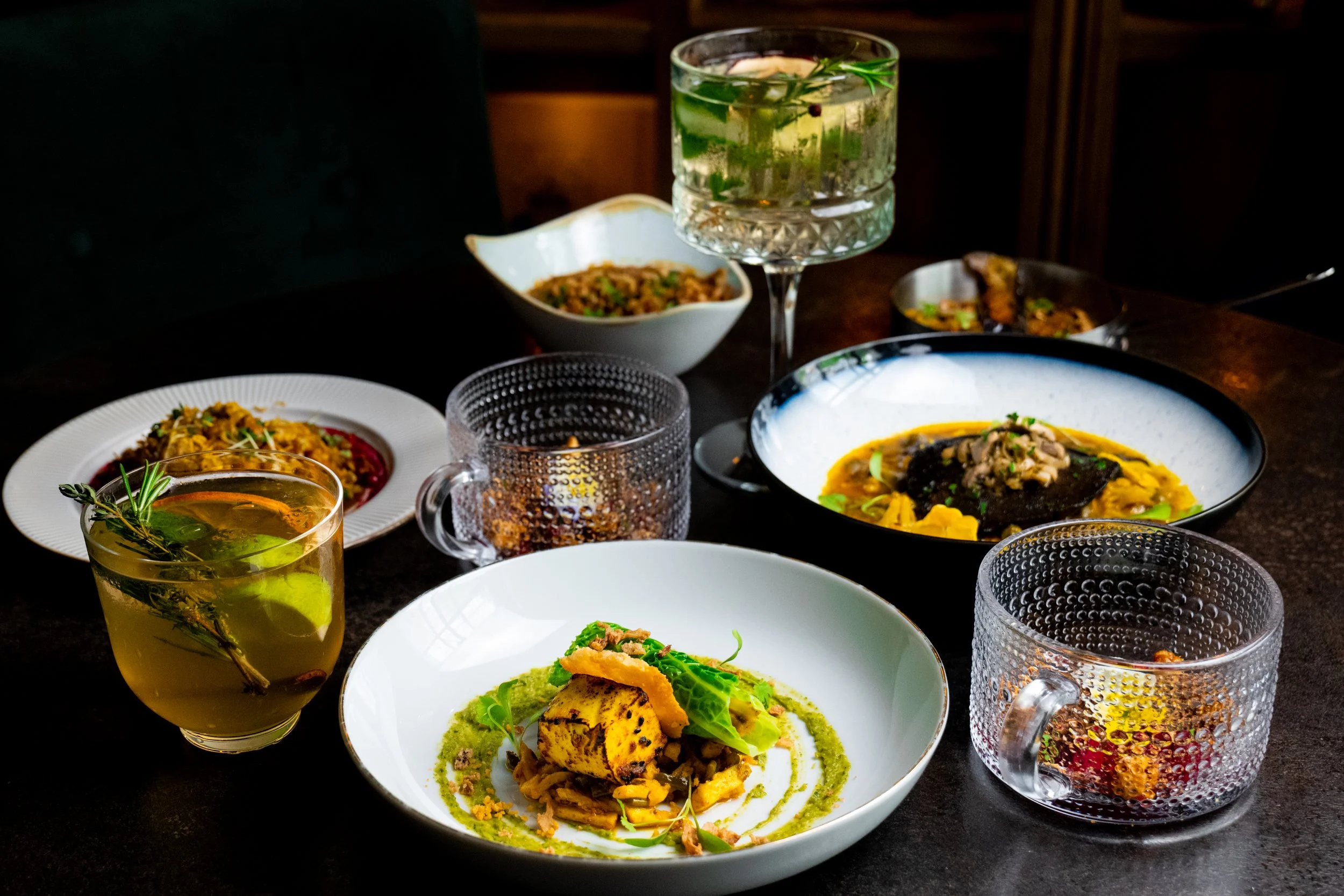 A table set with various dishes including a bowl of rice, a plate of grilled fish with mashed potatoes, and a salad, along with glasses of water and herbal drinks garnished with herbs.
