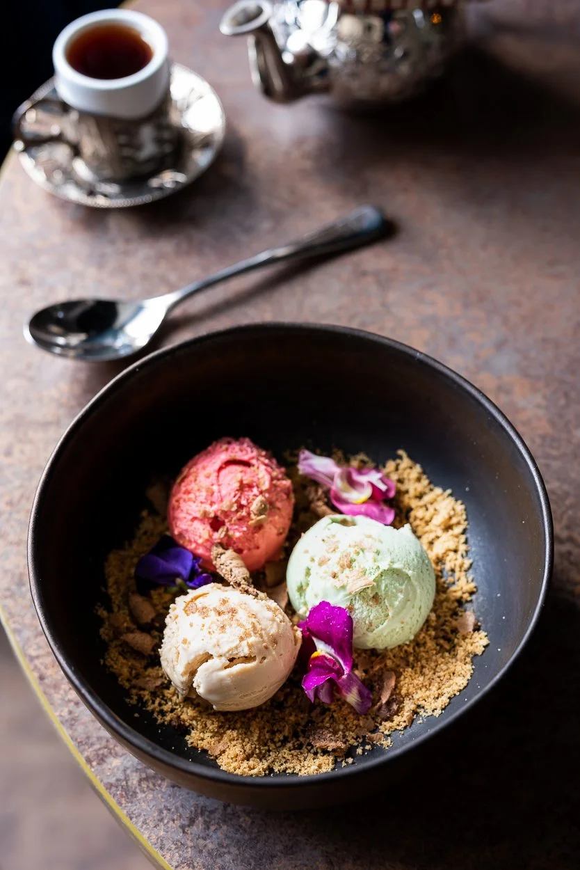 A black bowl of three scoops of ice cream with edible flower petals and crushed cookies on a brown table, with a spoon, a cup of tea, and a teapot in the background.