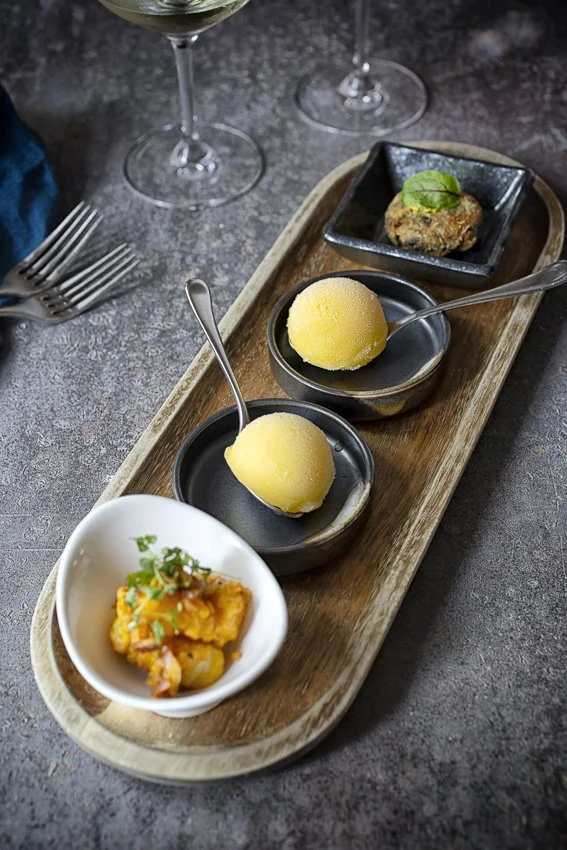Assorted appetizers on a wooden platter with wine glasses nearby.