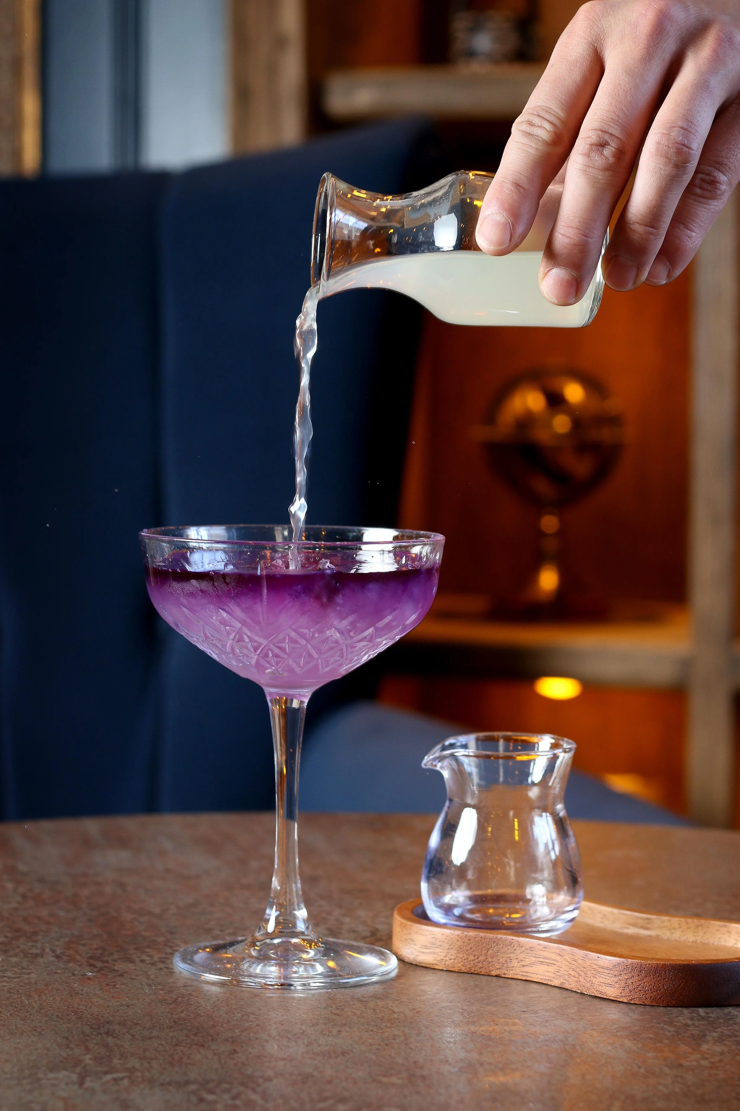 A hand pouring milk into a purple cocktail in a stemmed glass. The glass is on a wooden surface, with a small glass pitcher and a wooden tray nearby, in a cozy indoor setting.
