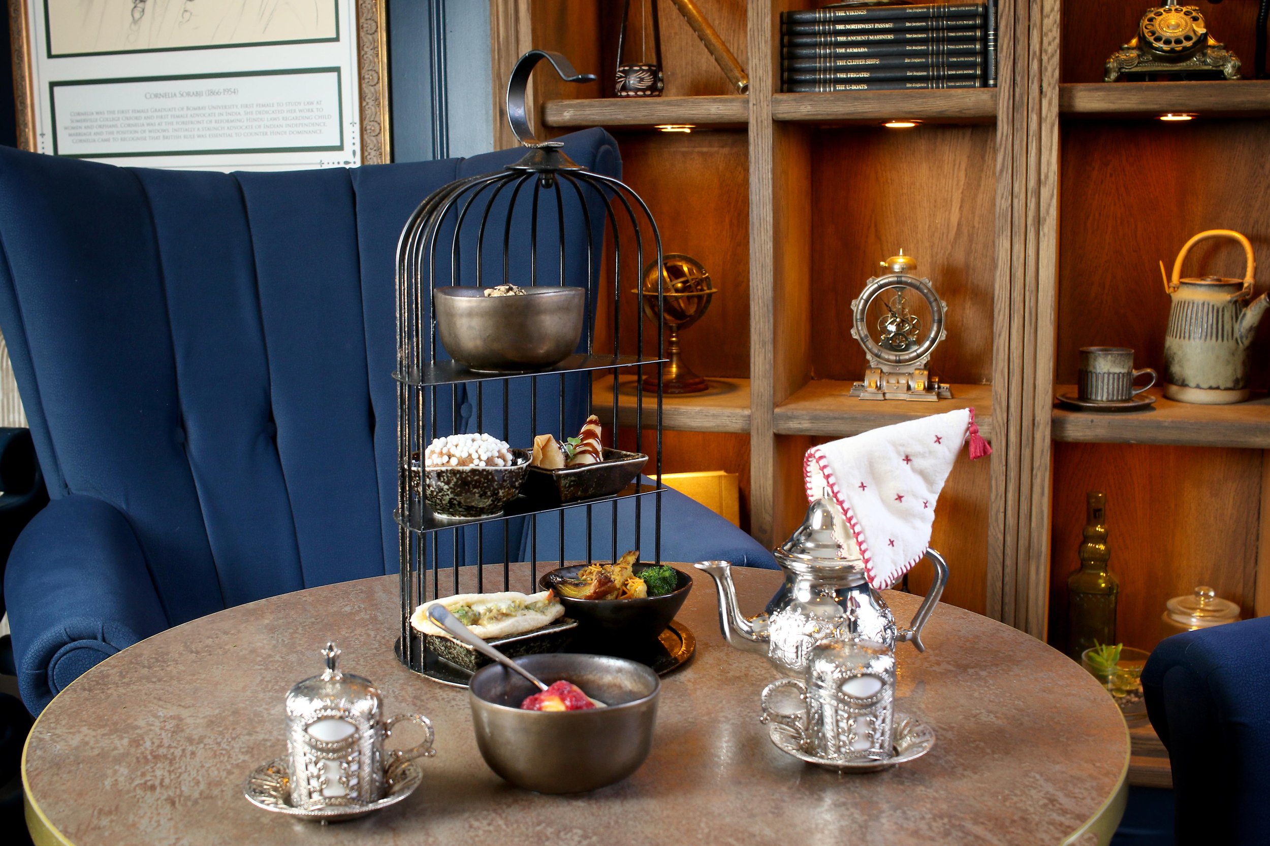 A table set for afternoon tea with a blue velvet armchair nearby. The table has a three-tiered stand with snacks, a silver teapot, and small candle holders. In the background, there is a wooden bookshelf with books and decorative items. The scene appears cozy and elegant.