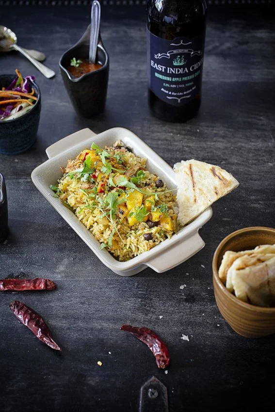 A plate of yellow rice with vegetables and herbs, a piece of flatbread, a small bowl of diced vegetables, a bottle of East India Creek drink, and some dried red chilies on a dark table.