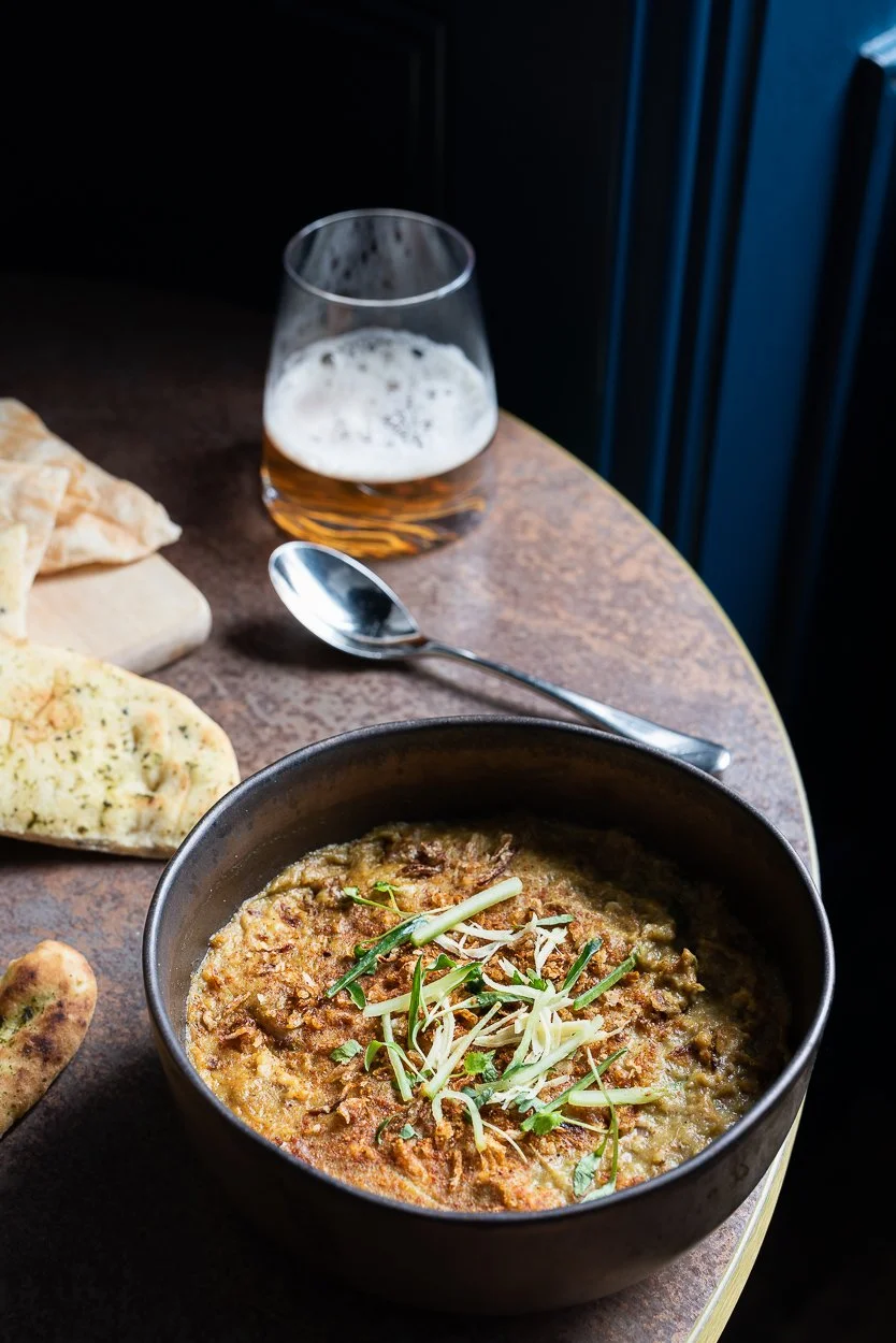 A bowl of Indian dish, possibly dal or curry, garnished with herbs, on a wooden table with naan bread, a spoon, and a glass of beer.