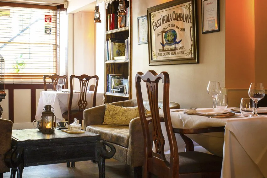 Cozy restaurant interior with tables set with wine glasses and plates, decorated with a vintage painting on the wall, a bookshelf filled with books, and a window with blinds letting in natural light.
