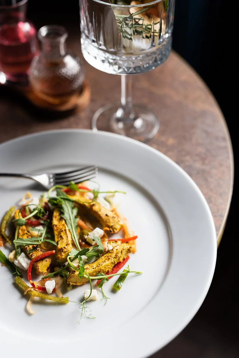A white plate of colorful vegetable stir-fry with sliced peppers, green beans, and microgreens on a wooden table. A glass of white wine and salt and pepper shakers are also visible.