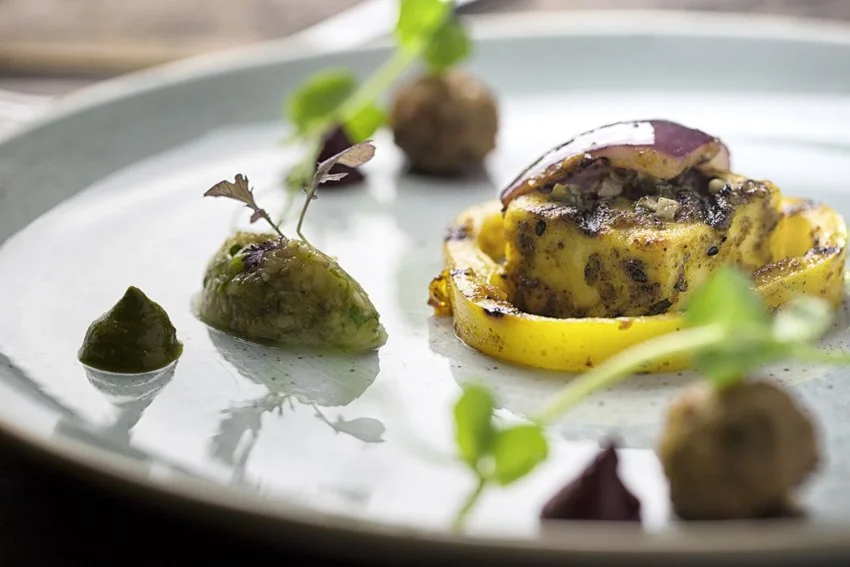 Close-up of a gourmet dish featuring a baked stuffed pepper with melted cheese and herbs, garnished with microgreens and small roasted vegetables on a white plate.