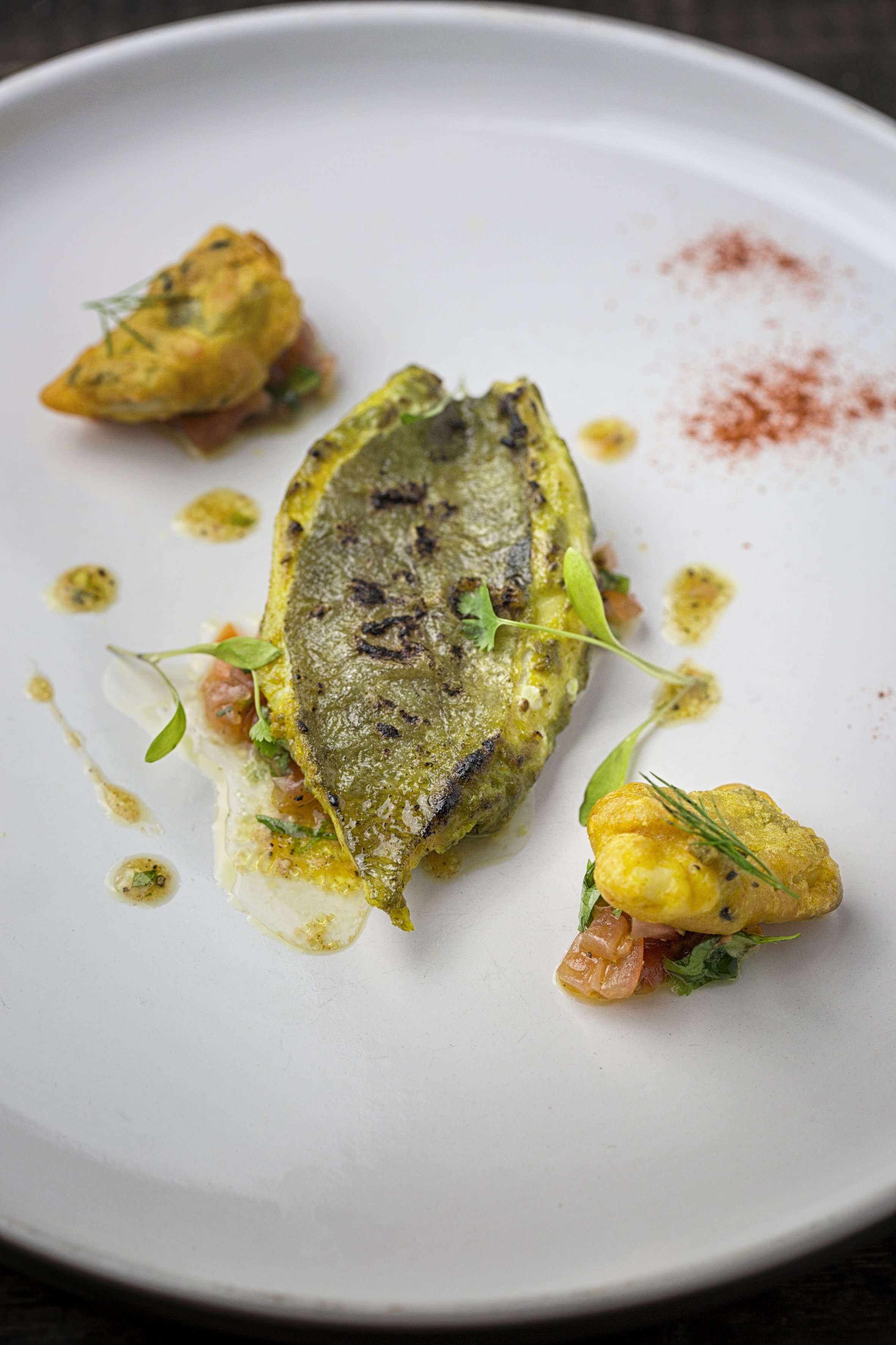 Close-up of a gourmet dish with a grilled fish fillet topped with a green sauce, garnished with microgreens, around it are small dollops of sauce, and two pieces of stuffed, fried plantains, on a white plate with a dark background.