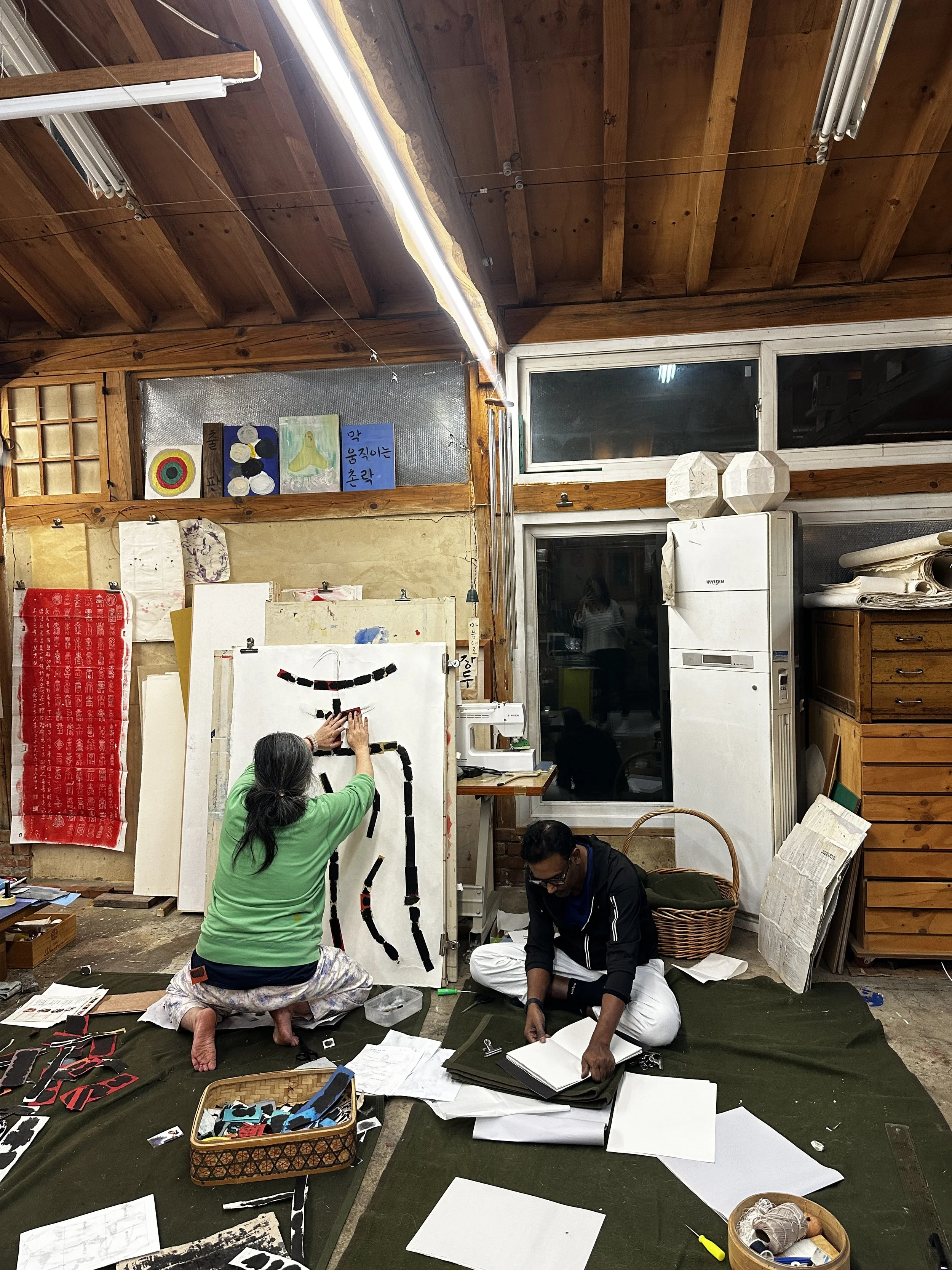 People working on an art project in a cozy, rustic indoor space with wooden ceiling beams. One person kneels on the floor, creating a large black and red artwork with paper, while another sits nearby, organizing papers and craft supplies.