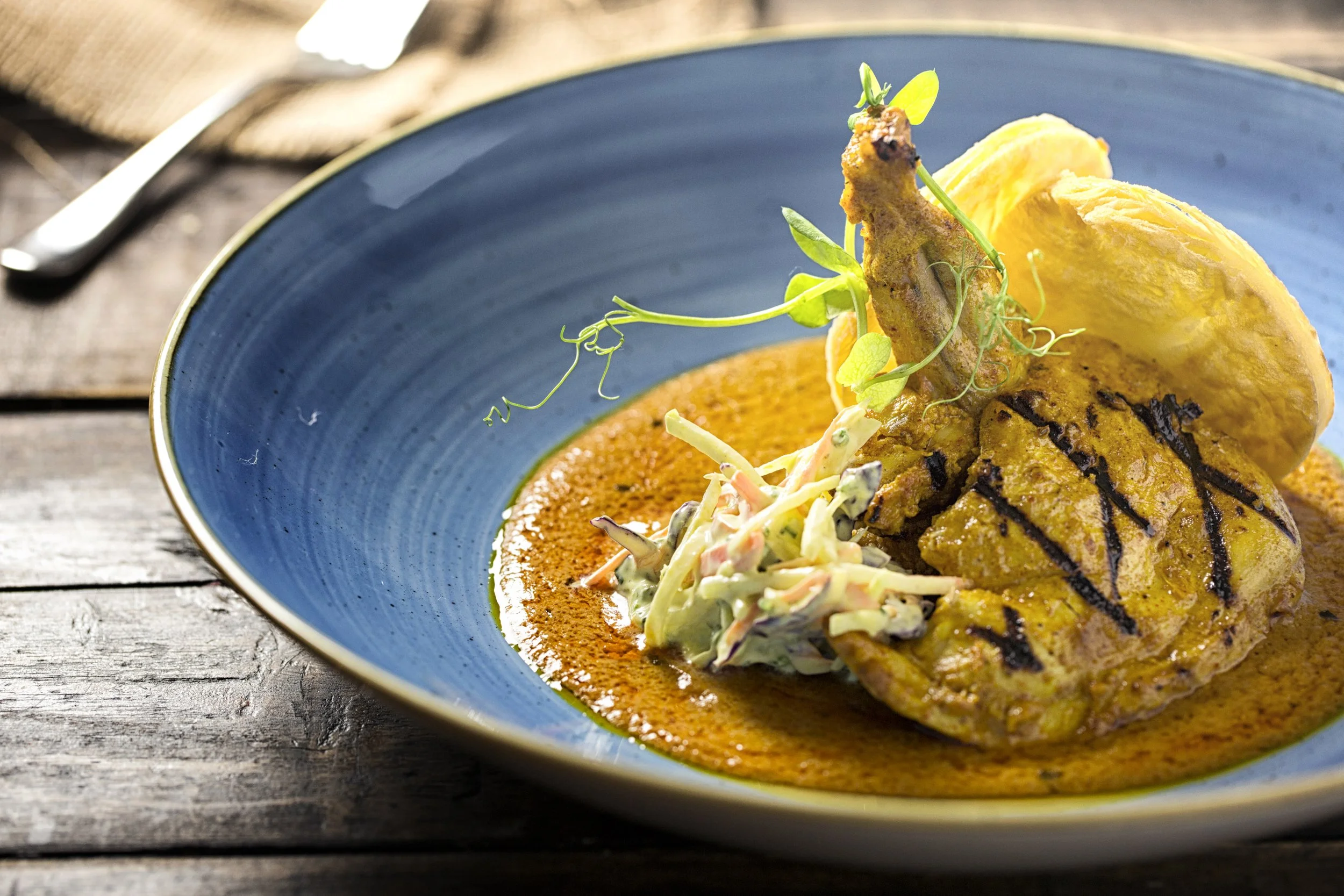 A blue bowl containing grilled chicken breast with grill marks, yellow potato chips, and a side of coleslaw, on a rustic wooden table.