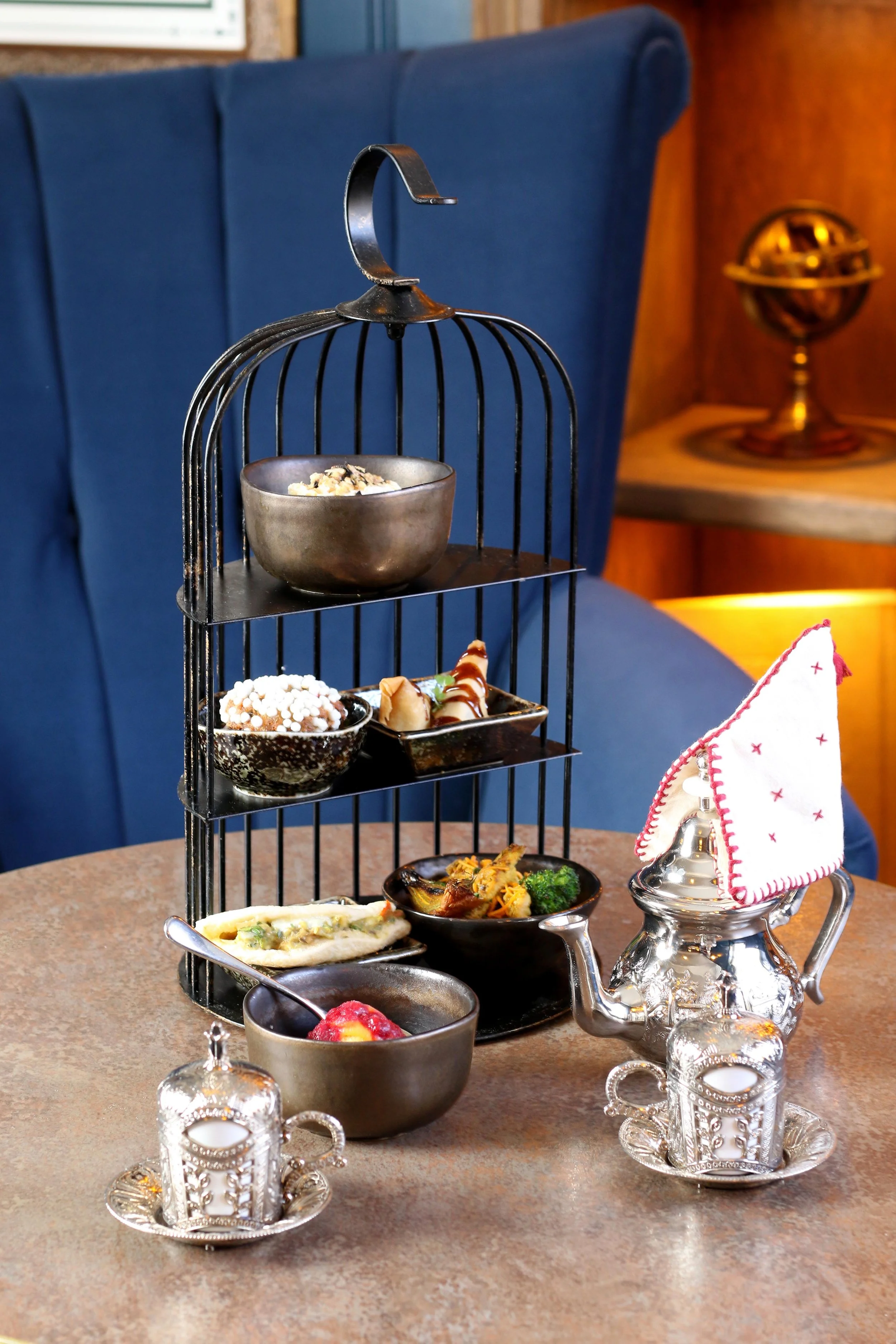Close-up of a multi-tiered metal serving stand with various foods, including a bowl of cereal or rice, cookies, a small sandwich, and a bowl of fruit, on a table with ornate silver candle holders and a glass teapot with a napkin, against a blue uphol