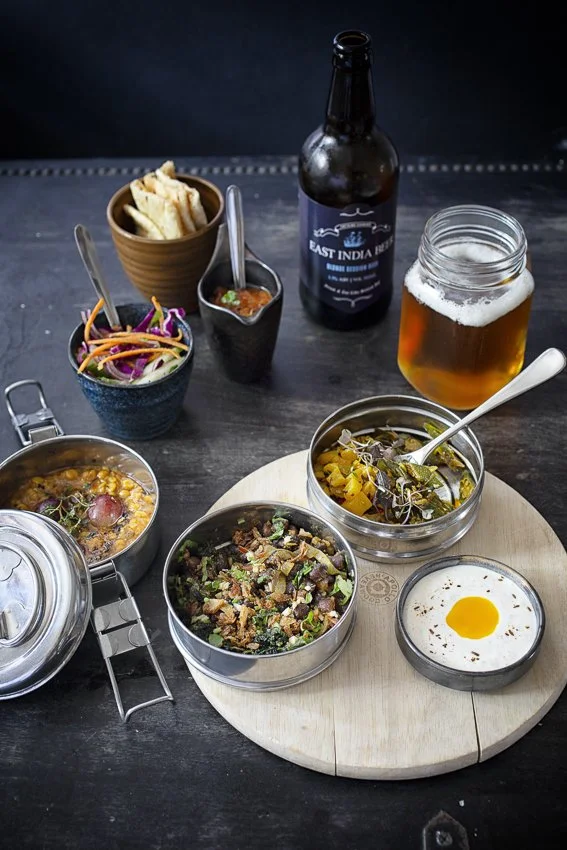 Indian cuisine meal with various dishes and drinks on a dark table. Includes bowls of curry, salad, fried egg, chips, a bottle of beer, and a mason jar of beer.