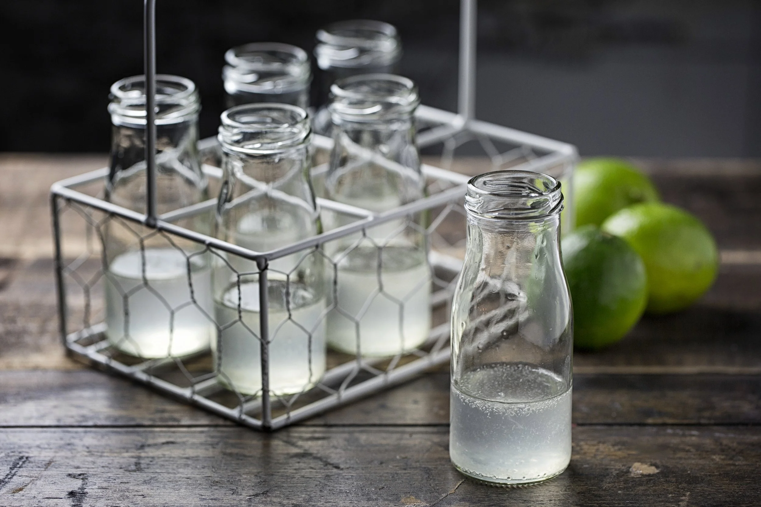 Several small glass bottles filled with a clear liquid are arranged in a white wire rack on a wooden surface. Two green apples are in the background.