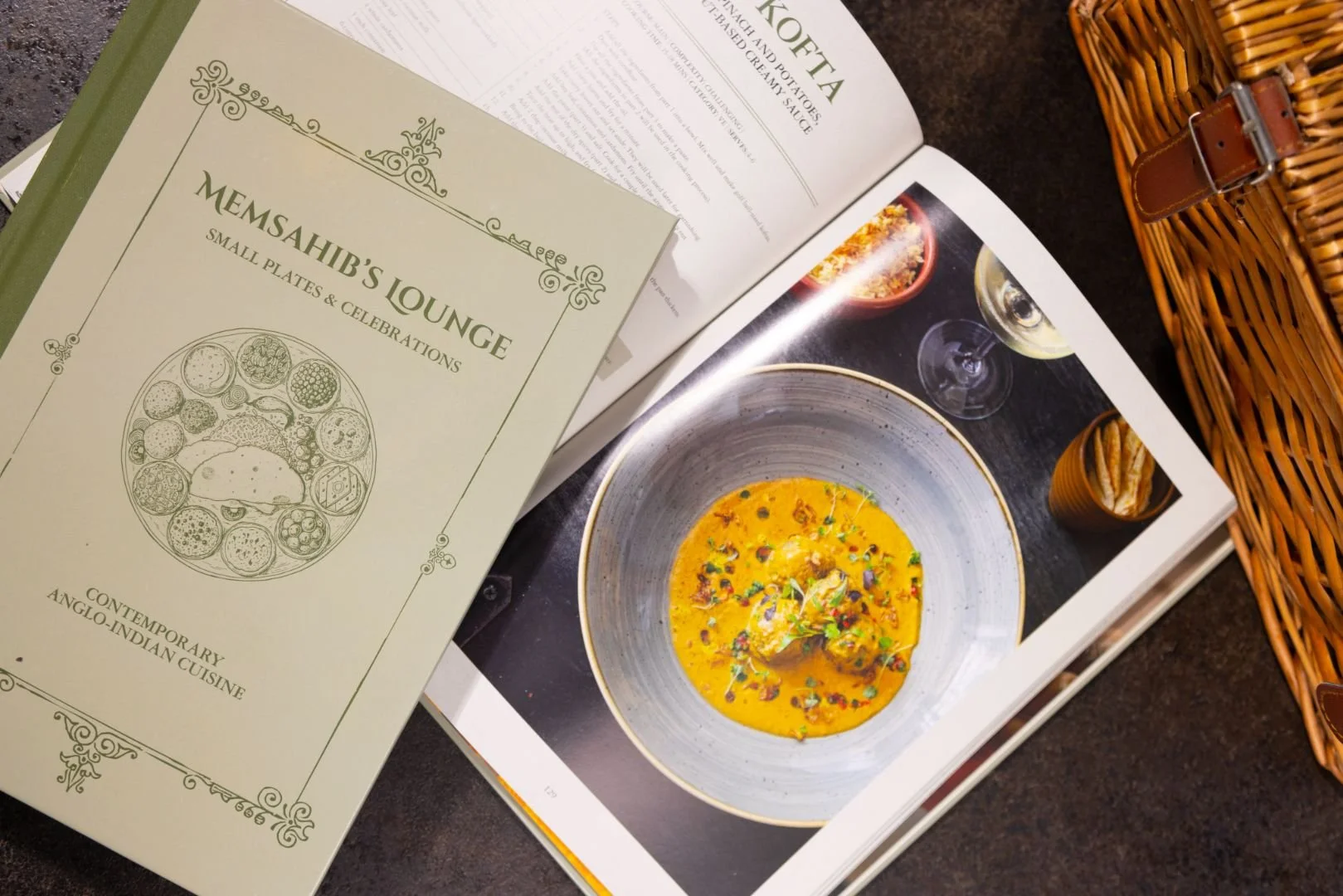 A menu booklet labeled 'Memsaheb's Lounge Small Plates & Celebrations' featuring an illustration of various small dishes and labeled as contemporary Anglo-Indian cuisine. Next to the menu is an open cookbook displaying a photograph of a yellow curry in a bowl, garnished with herbs, alongside a glass of white wine and a small bowl of crispy snacks, with a wicker basket nearby on a dark surface.
