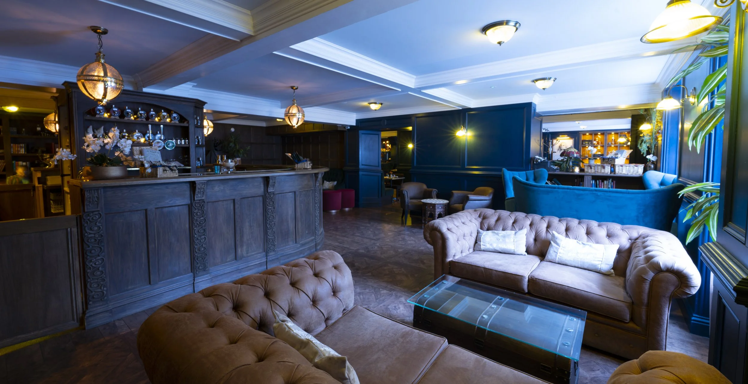 A cozy lounge with plush leather armchairs, a glass coffee table, and a bar area with wooden cabinets, decorative lighting, and plants.