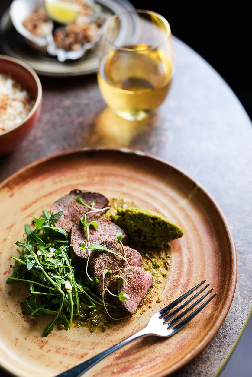 A plate with cooked meat, garnished with microgreens, accompanied by a slice of green vegetable cake or bread, with a glass of white wine in the background and bowls of other dishes on a dark table.