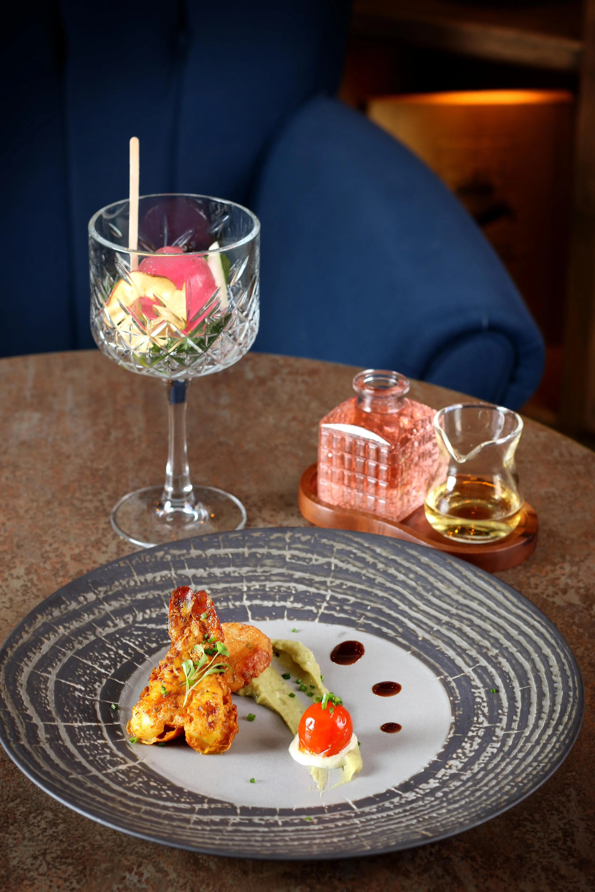 A crystal glass with a pink flower and green leaves, containing a drink with a wooden stir stick, on a wooden table. In the background, a pink salt lamp, small glass pitcher with a yellow liquid, and another glass container are visible. Part of a pla