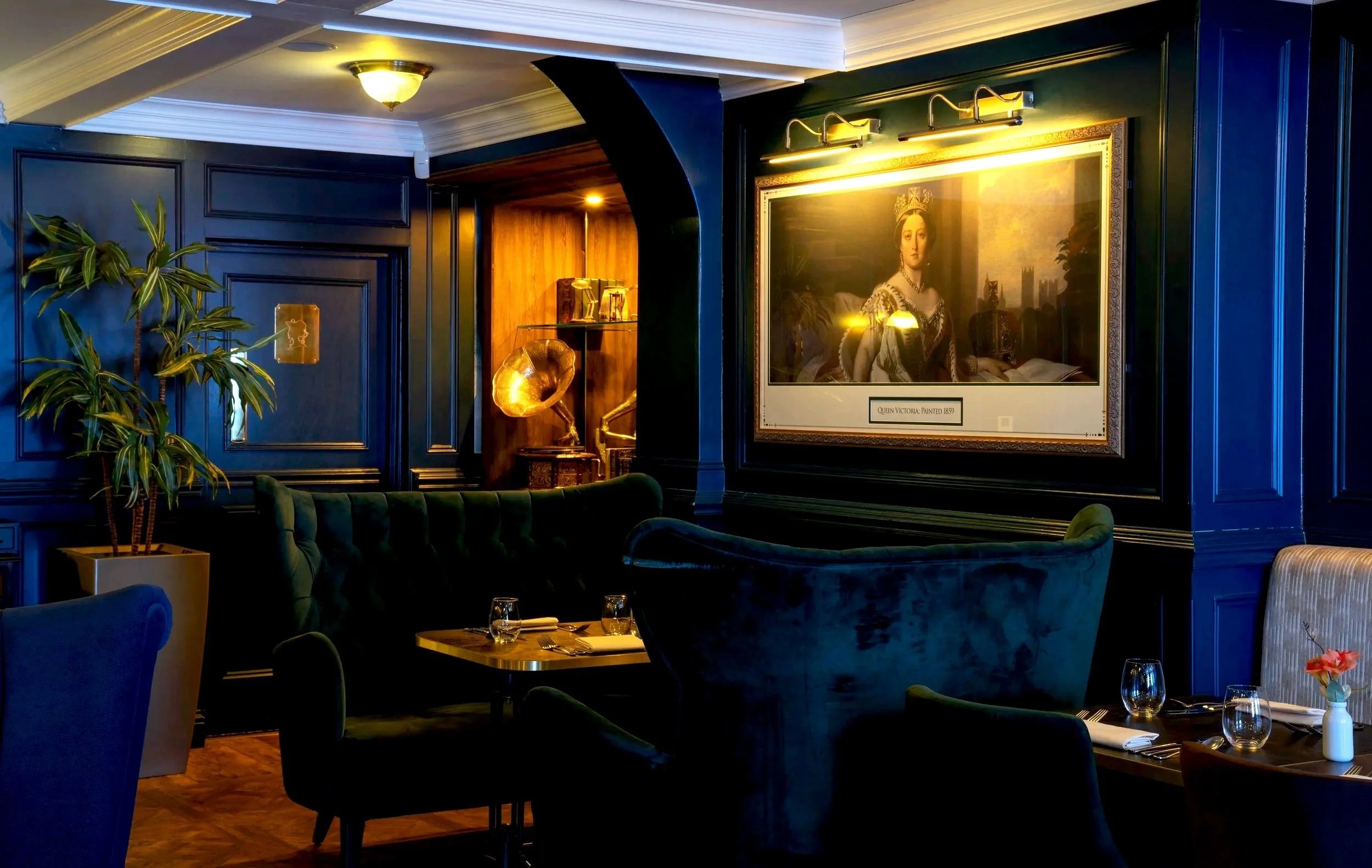 A dimly lit upscale restaurant with dark blue paneled walls, elegant velvet chairs, and a framed portrait of Queen Victoria on the wall. The table is set with wine glasses, napkins, and a small flower arrangement.