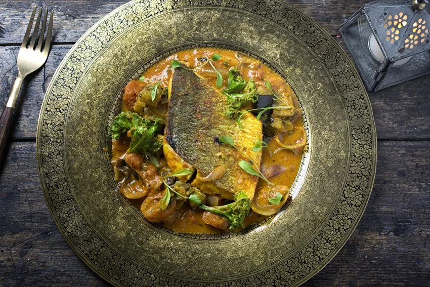 Plate with cooked fish fillet, vegetables, and sauce on a decorative gold-rimmed plate, placed on a dark wooden table. A fork is on the left side, and a lantern is on the right.