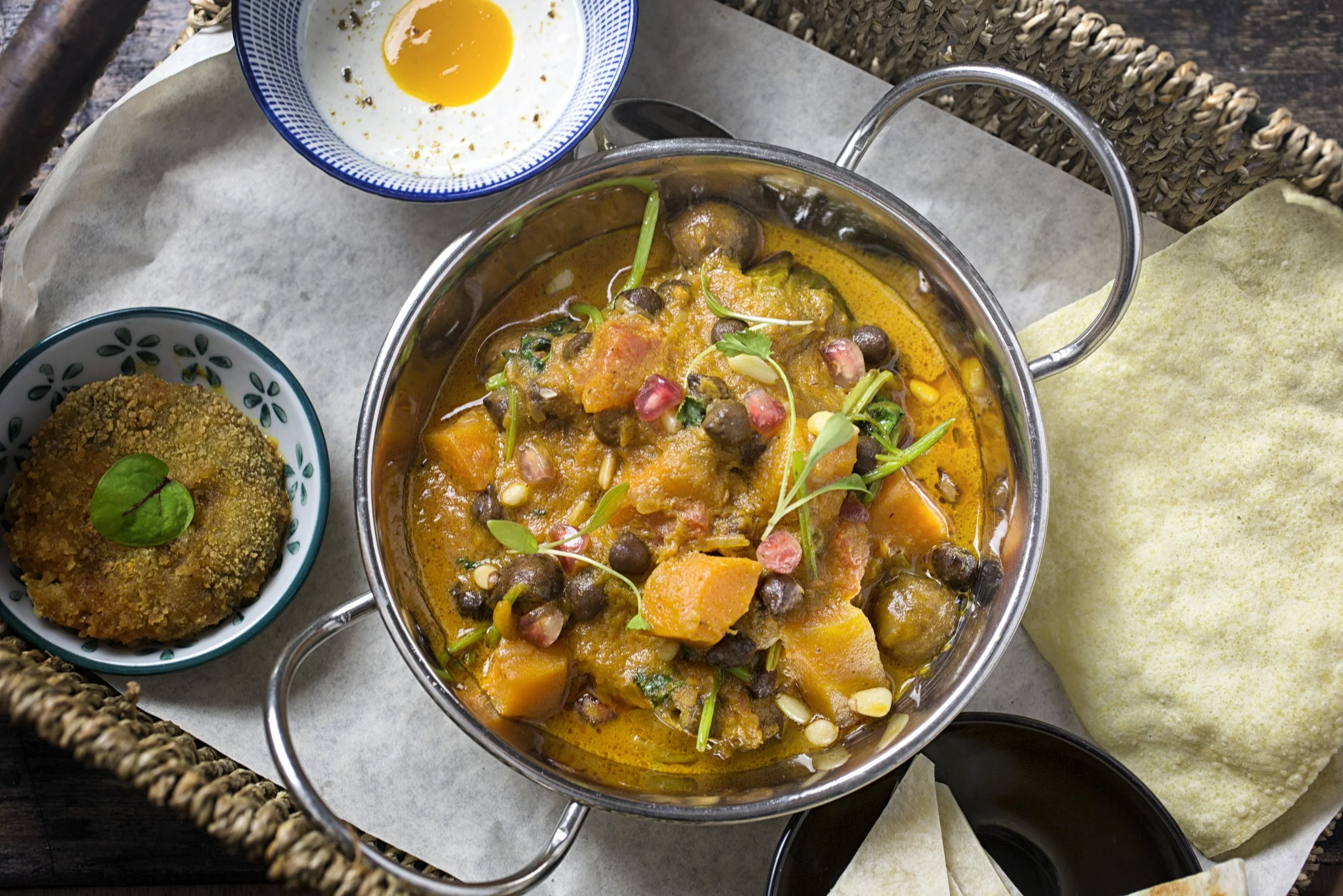 A pot of Indian curry with sweet potatoes, chickpeas, and chopped vegetables, garnished with microgreens, alongside a bowl of fried snack and a bowl of a boiled egg. The food is placed on a woven tray with parchment paper.