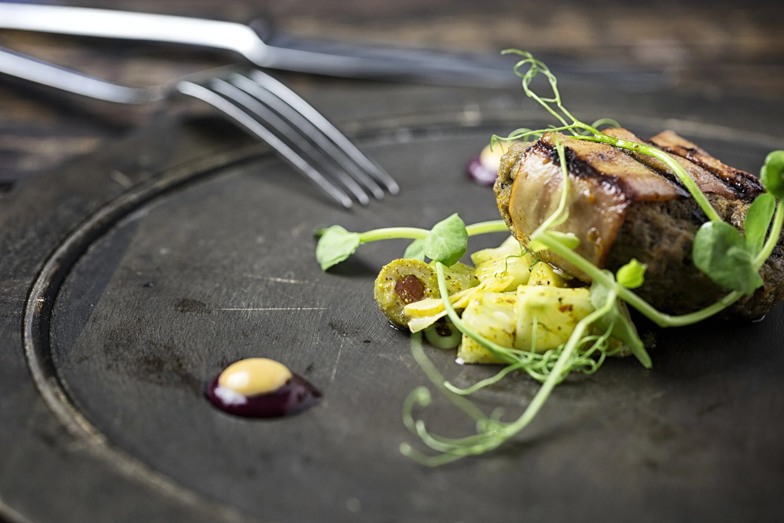 Close-up of a plated gourmet dish with a piece of glazed meat, pasta, microgreens, and a drizzle of sauce, on a dark serving plate with silverware in the background.